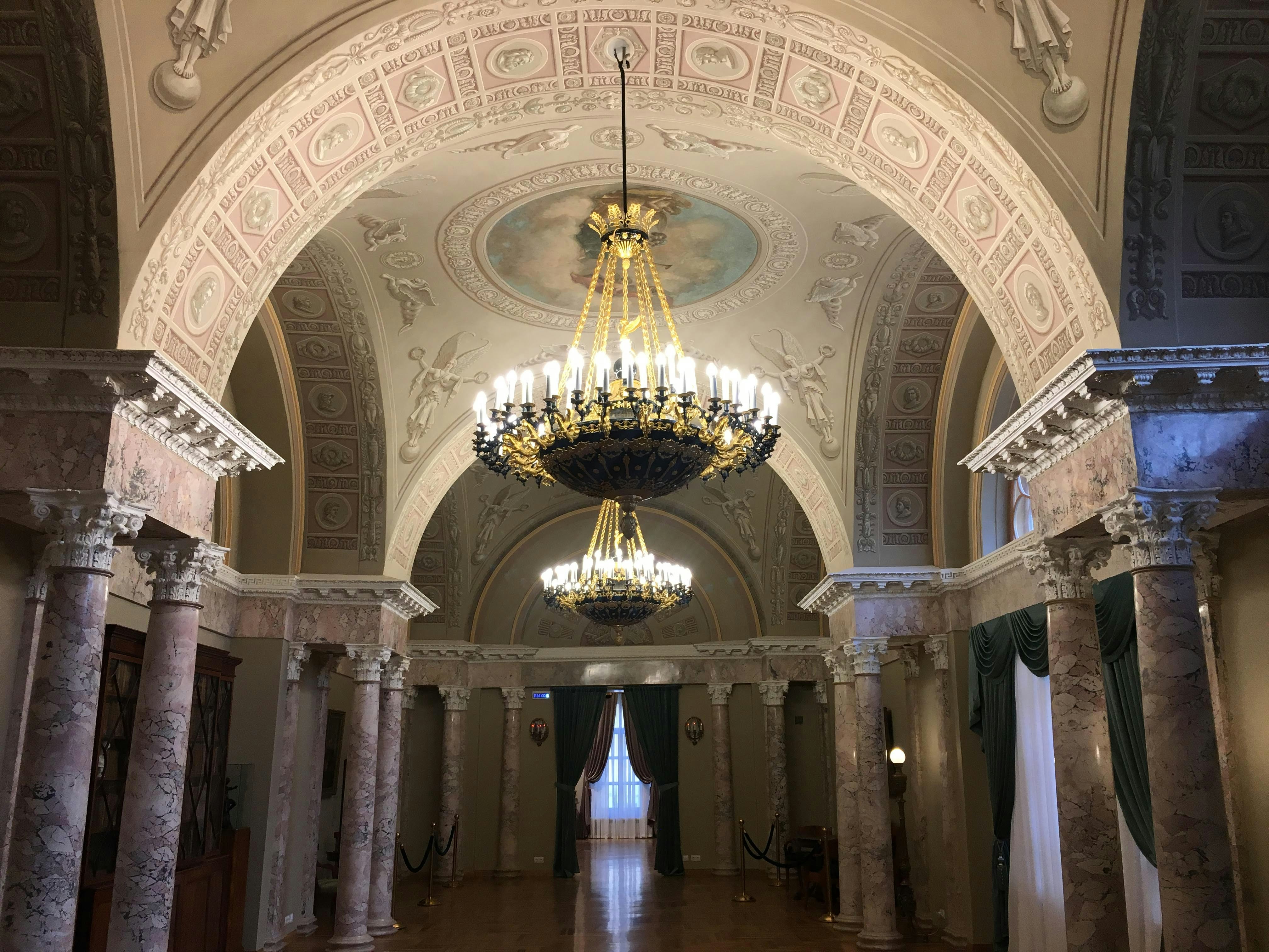 A grand hallway lined with marble columns, with an arched ceiling covered in paintings and motifs. Two large chandeliers hang down from the vaulted ceiling. 