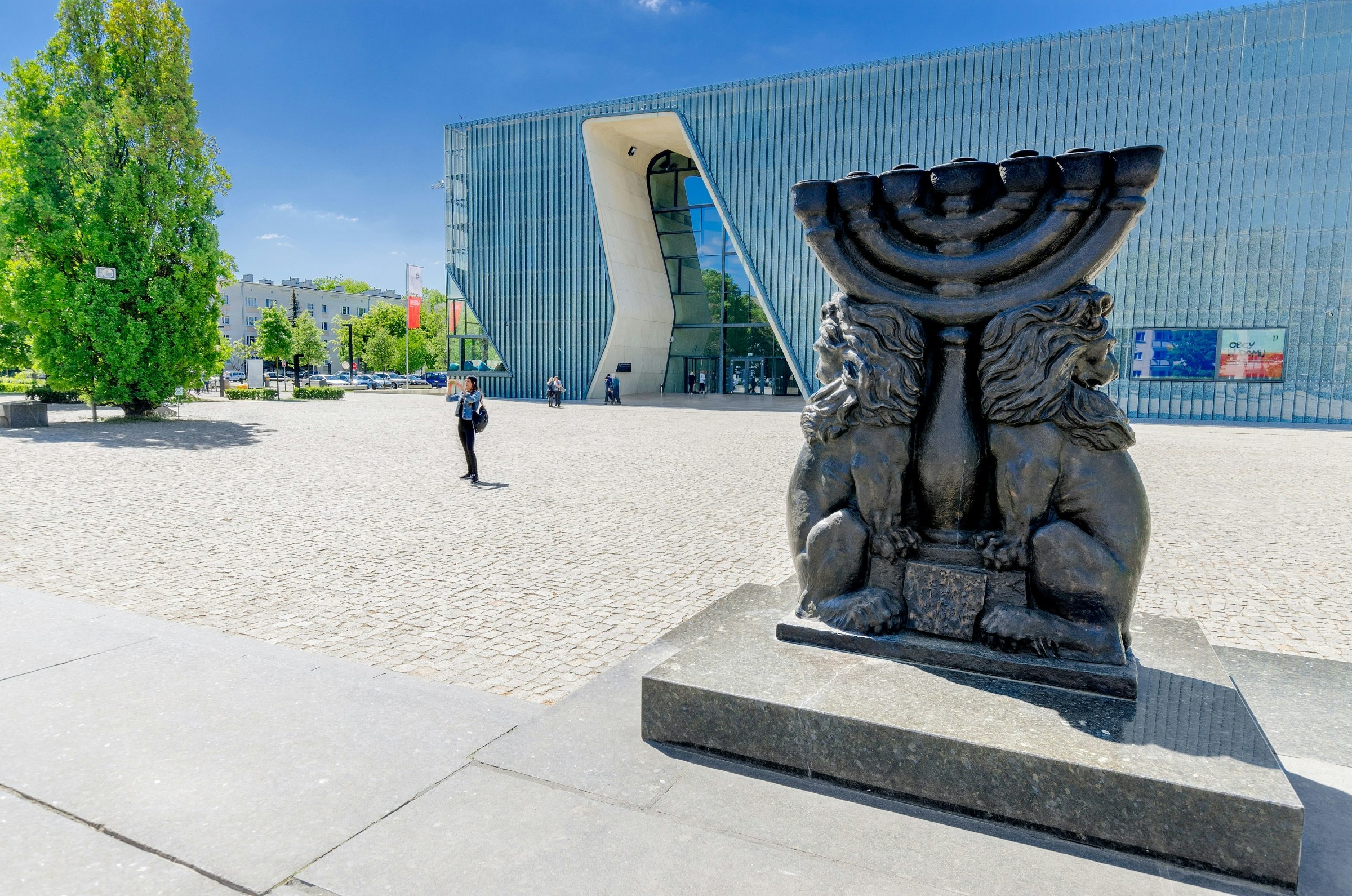 A sculpture of a menorah stands in an open square. A contemporary glass-and-steel museum building stands behind