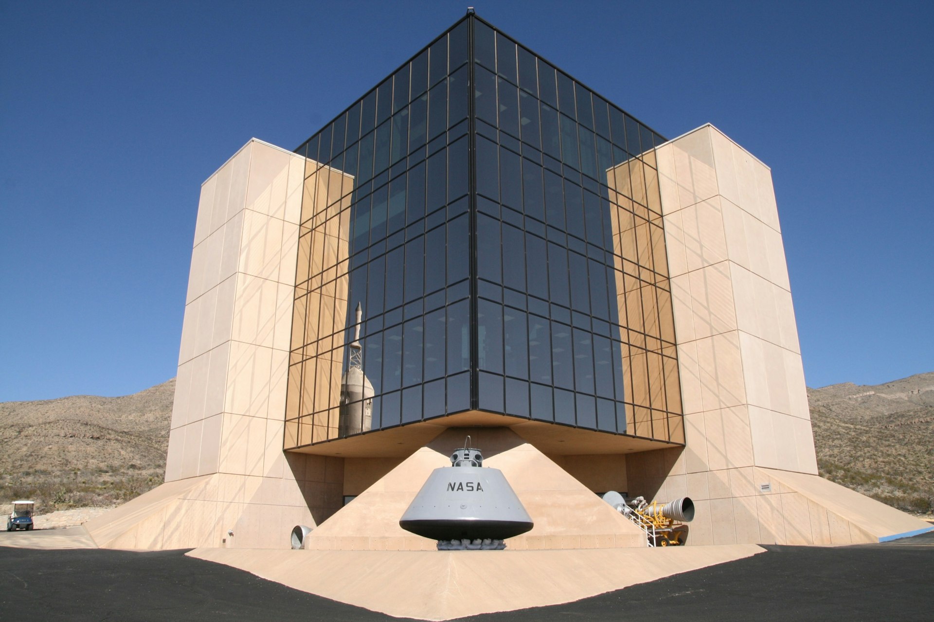 A modern glass building appears to be hovering between concrete pillars, with space equipments placed around and reflected in the windows