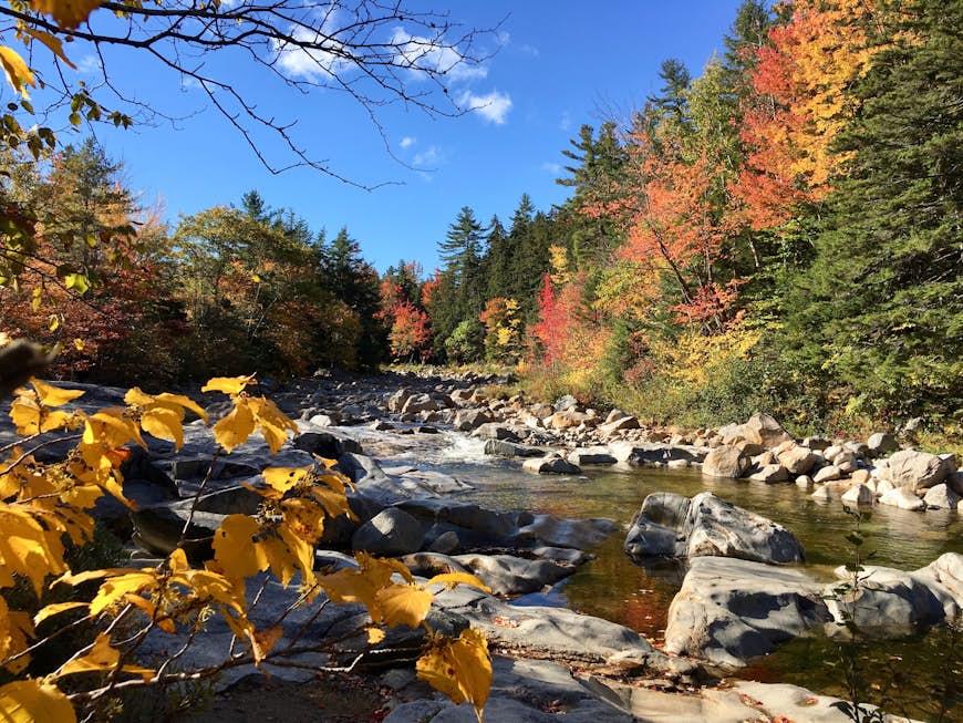 A stream tumbles over gray boulders as fall leaves stick out all around; New England fall foliage road trip