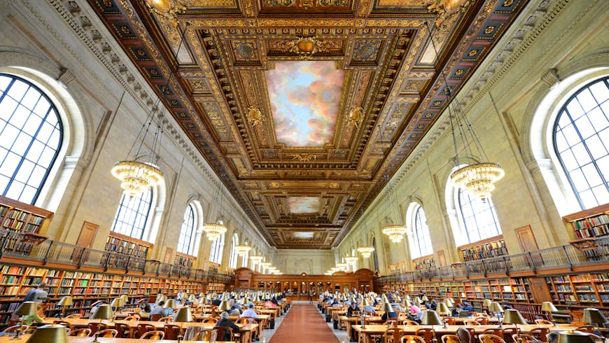 Interior shot of the expansive Rose Main Reading room at the New York Public Library.  There are multiple wooden tables and a very ornate ceiling with large chandeliers.