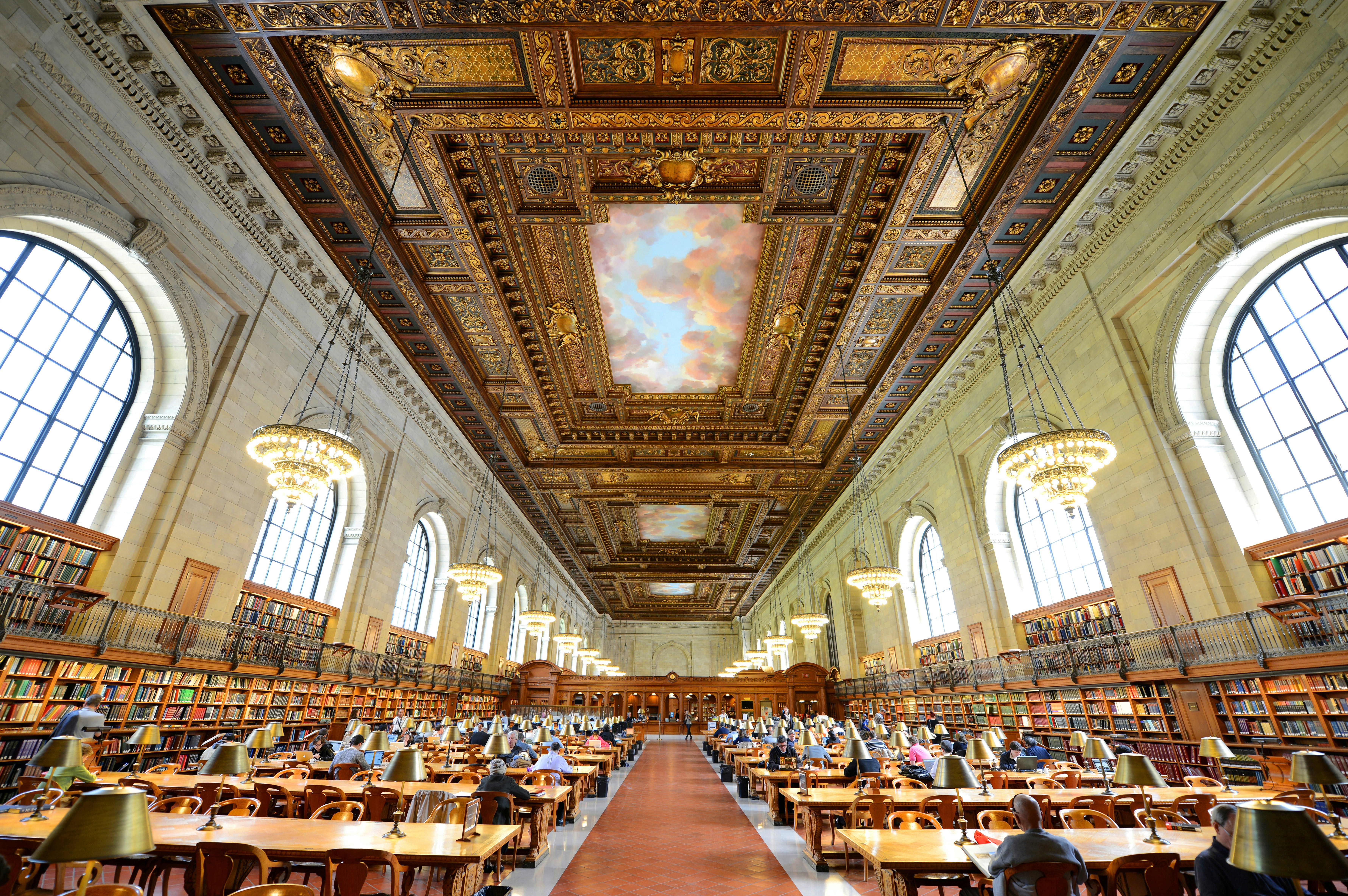 Interior shot of the expansive Rose Main Reading room at the New York Public Library. There are multiple wooden tables and a very ornate ceiling with large chandeliers.