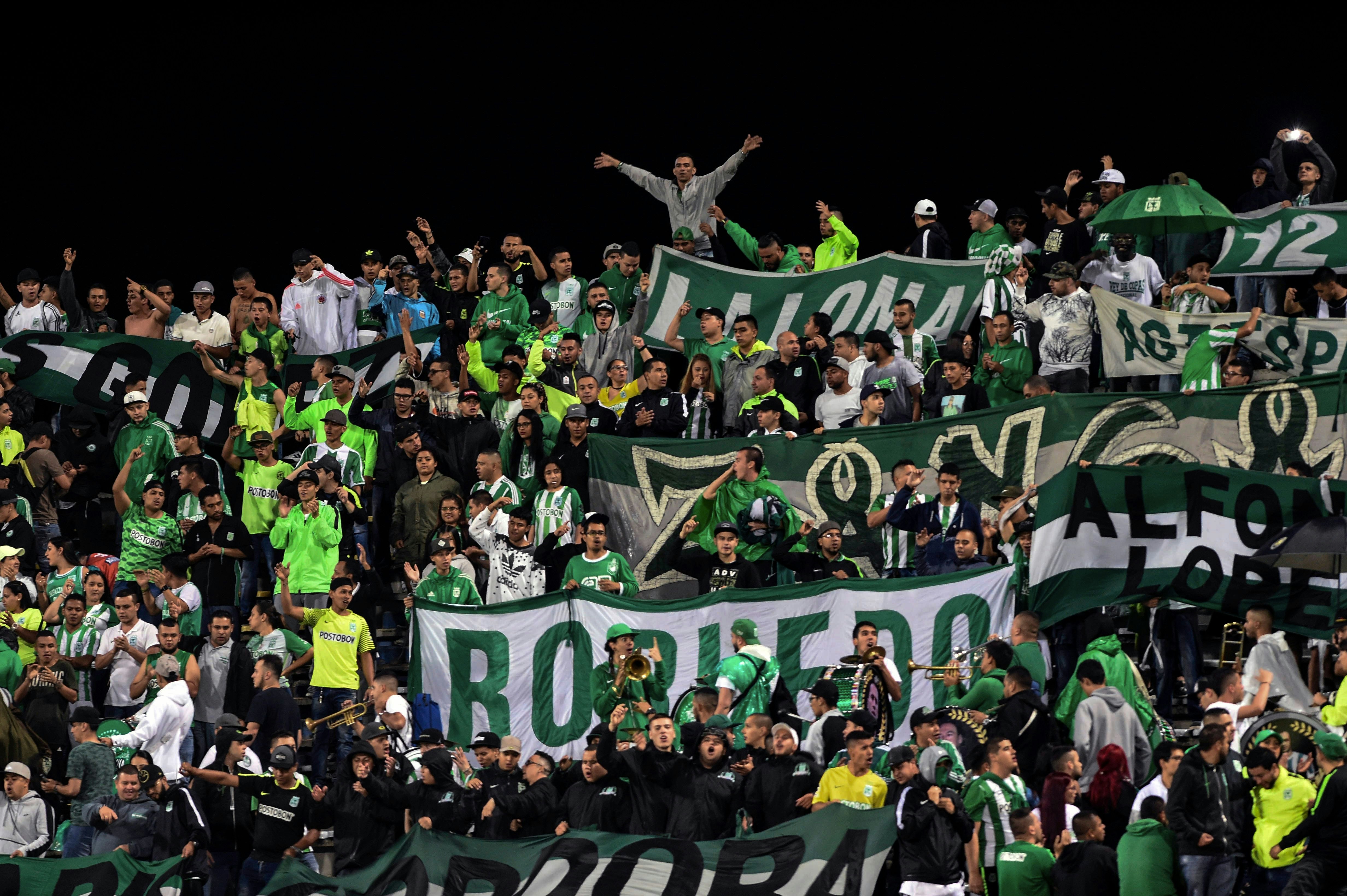 A large group of fans decked out in green and white wave flags and large banners in a stadium in Colombia.