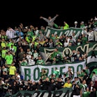 A large group of fans dressed in green and white wave flags and large banners in a stadium in Colombia.