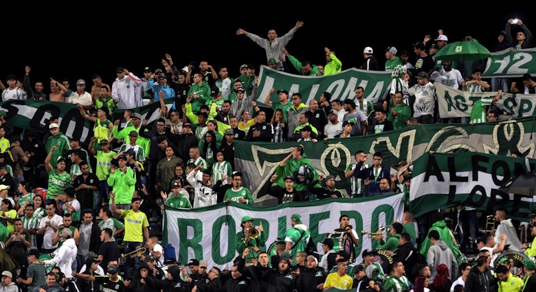 A large group of fans dressed in green and white wave flags and large banners in a stadium in Colombia.