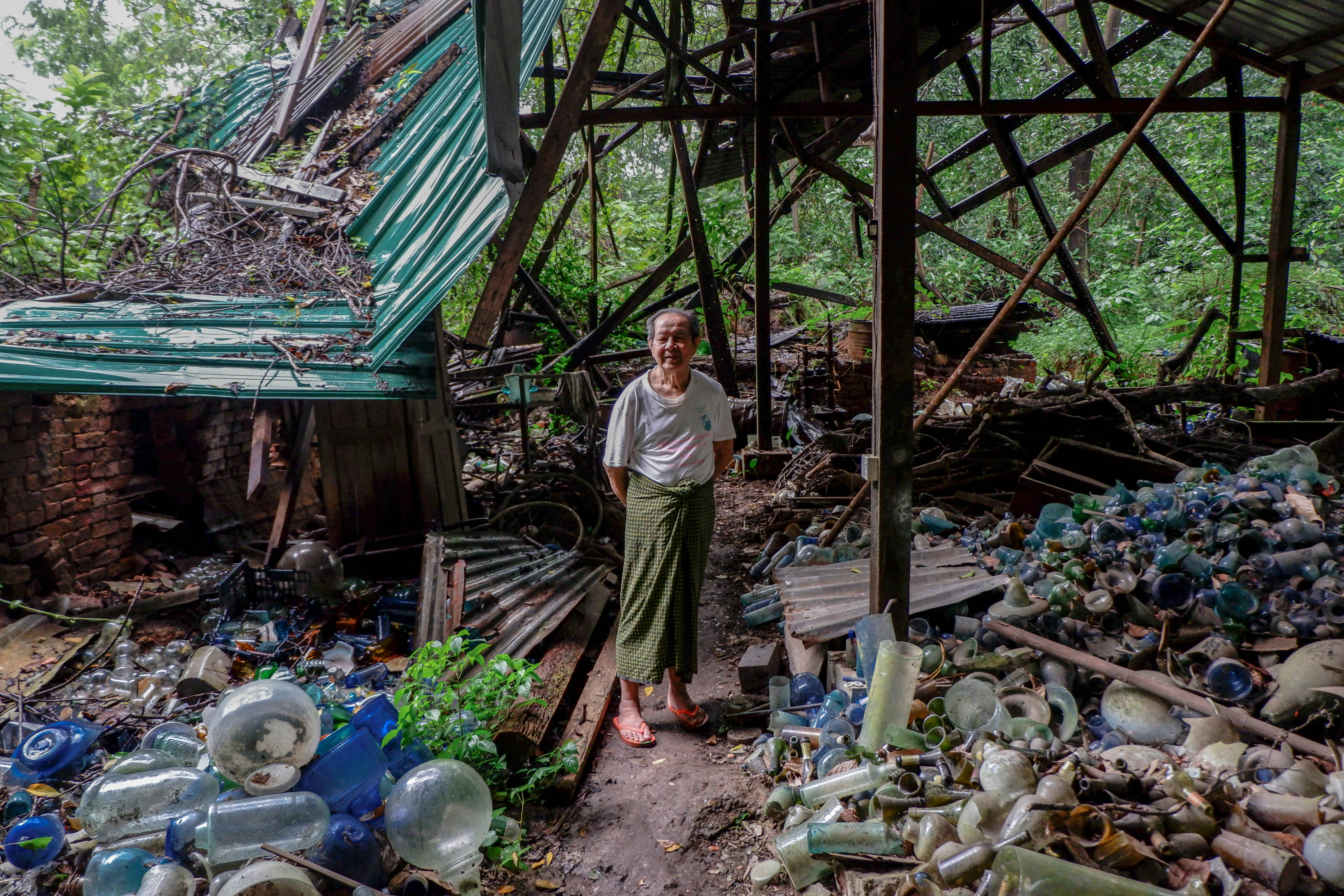 Thein Zaw, wearing a green 'longyi', white t-shirt and sandals, stands amongst the glass rubble at Nagar Glass Factory, which was destroyed by a cyclone.