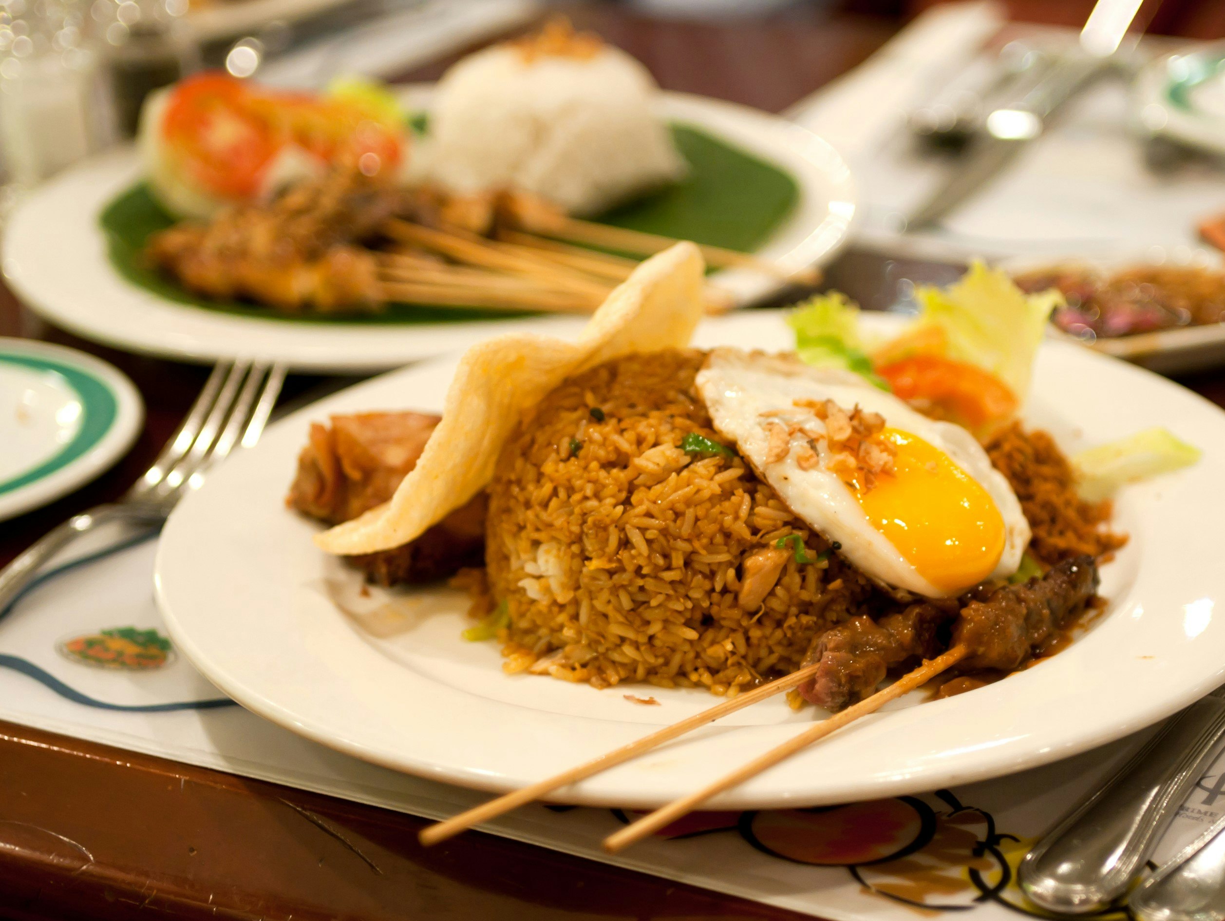 A plate of nasi goreng on a table. The dish consists primarily of rice and vegetables, topped with an egg. In this instance the dish is paired with chicken skewers.