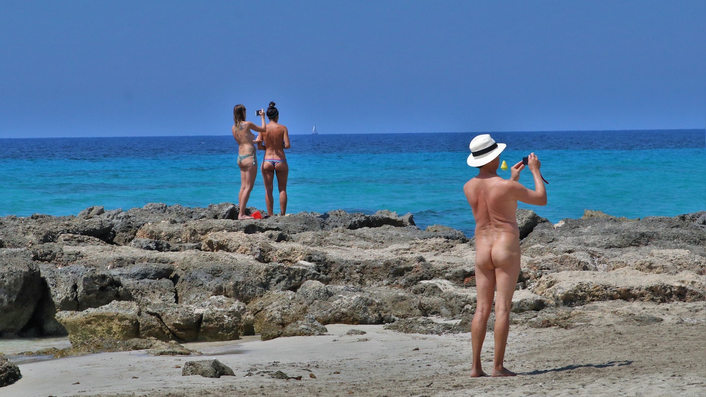A man wearing a Panama hat and nothing else, takes a photo of the sea. In the background are a pair of topless women also taking photos of the ocean