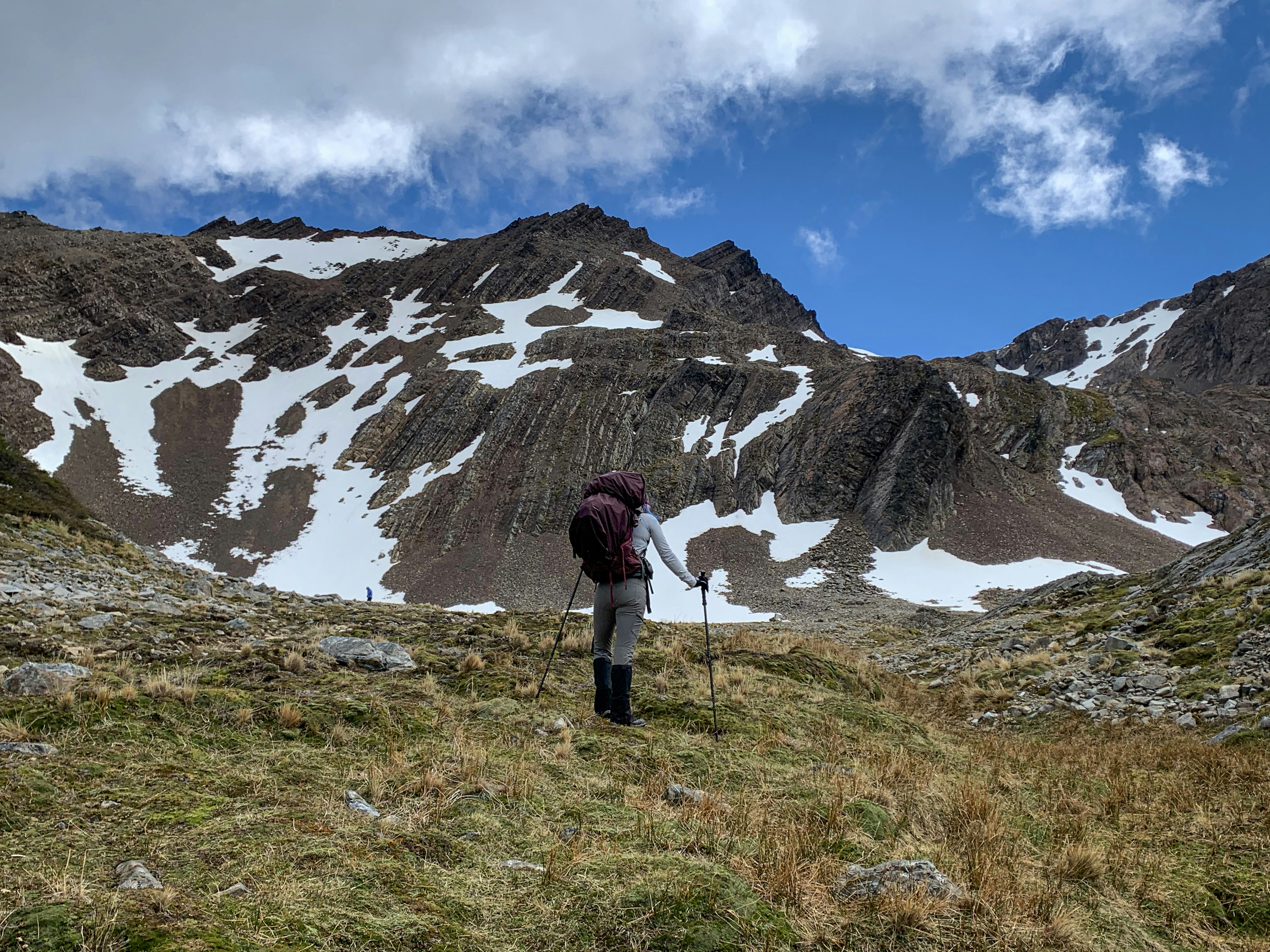 Hiker with backpack and hiking poles stands on a grassy hill looking at snow-streaked mountains