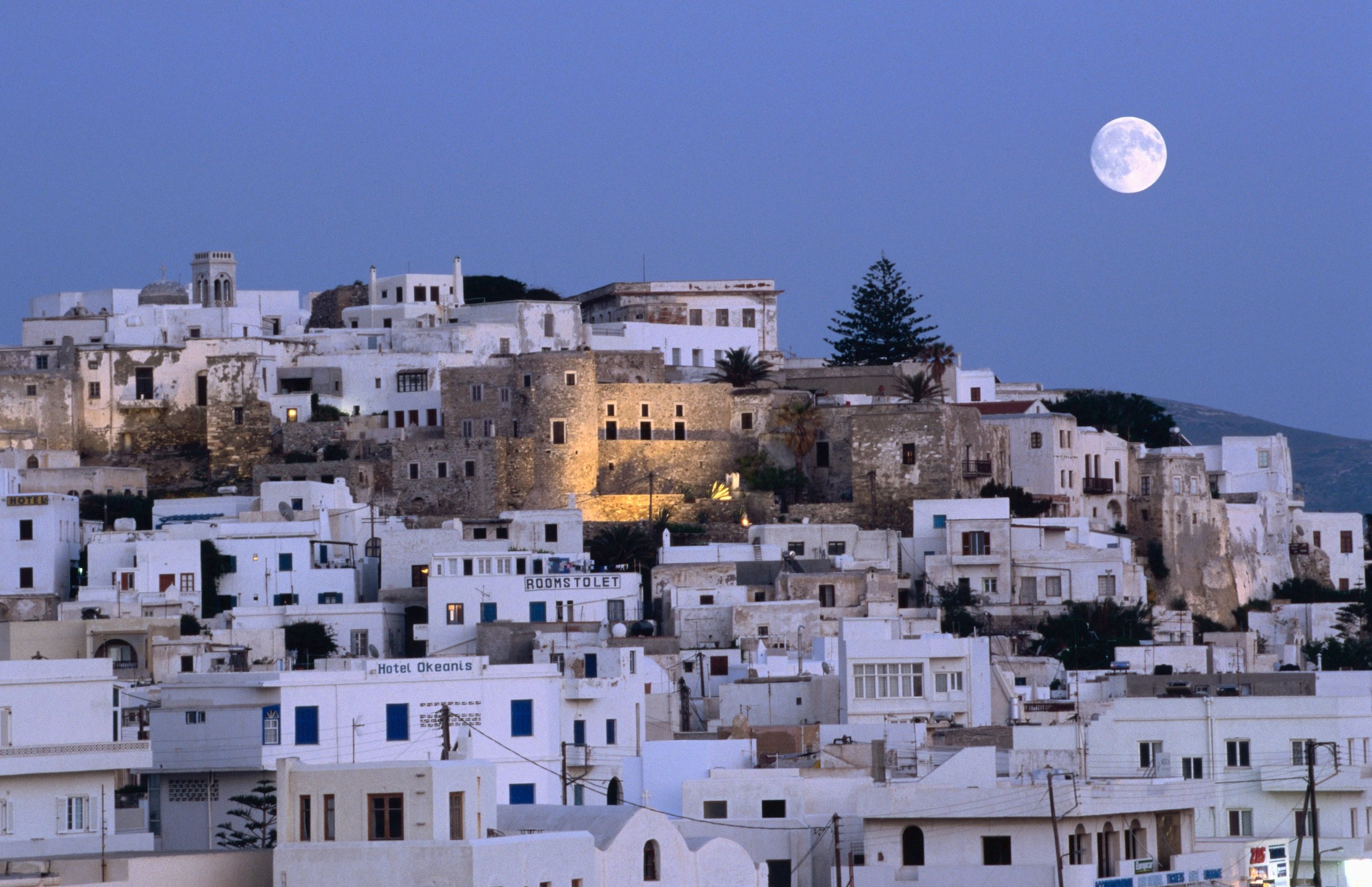 A full moon over a town of boxy white buildings packed together on a hillside. A castle structure sits in the middle.