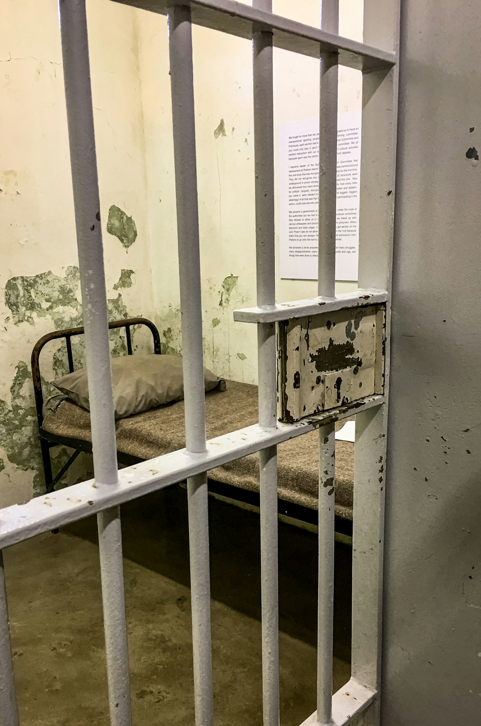 A small bed is visible within a tiny cell through the barred prison door.