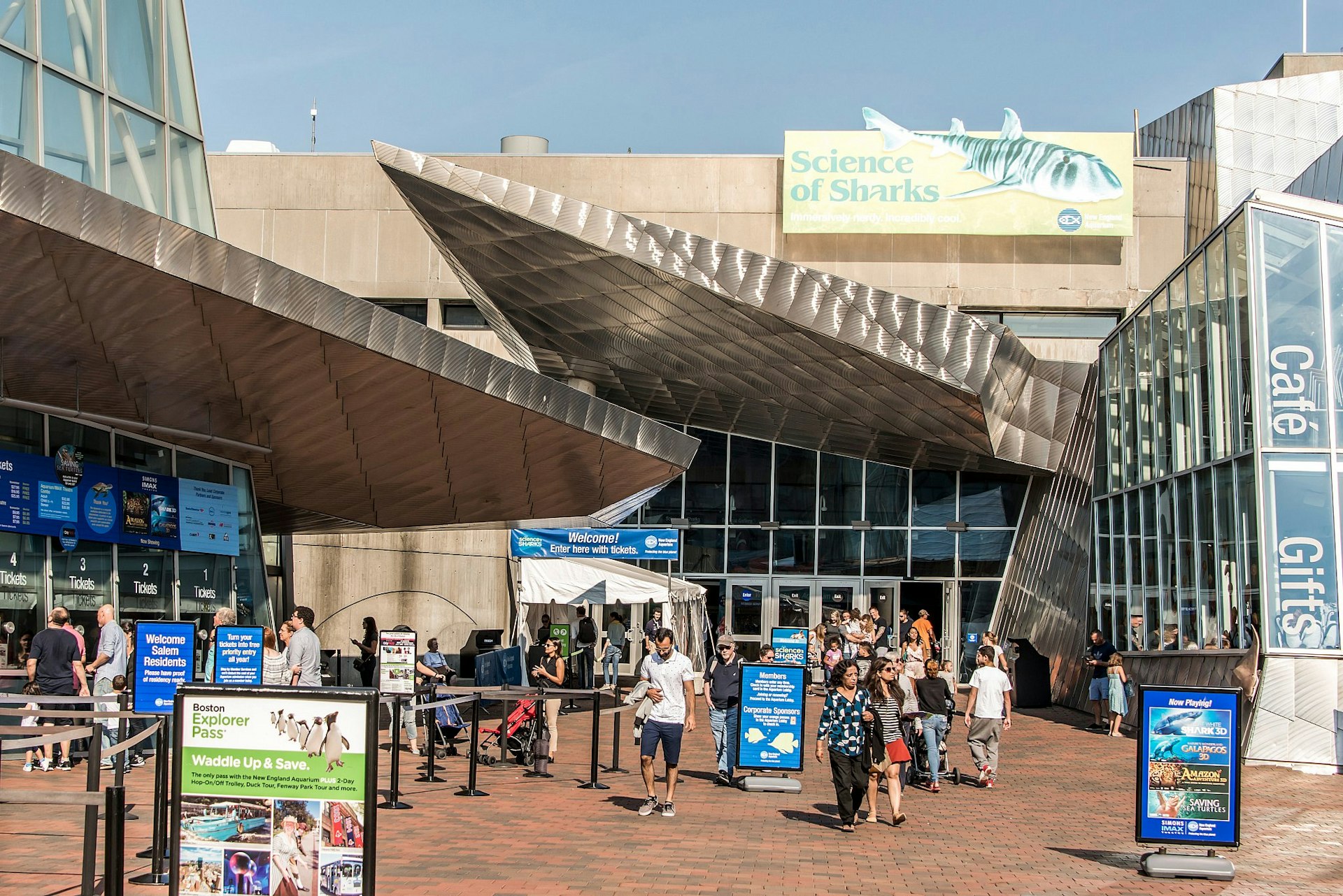 The exterior of the New England Aquarium, Boston; it's a concrete and glass building with a jagged metal canopy that resembles shark's fins; crowds mill about outside. 
