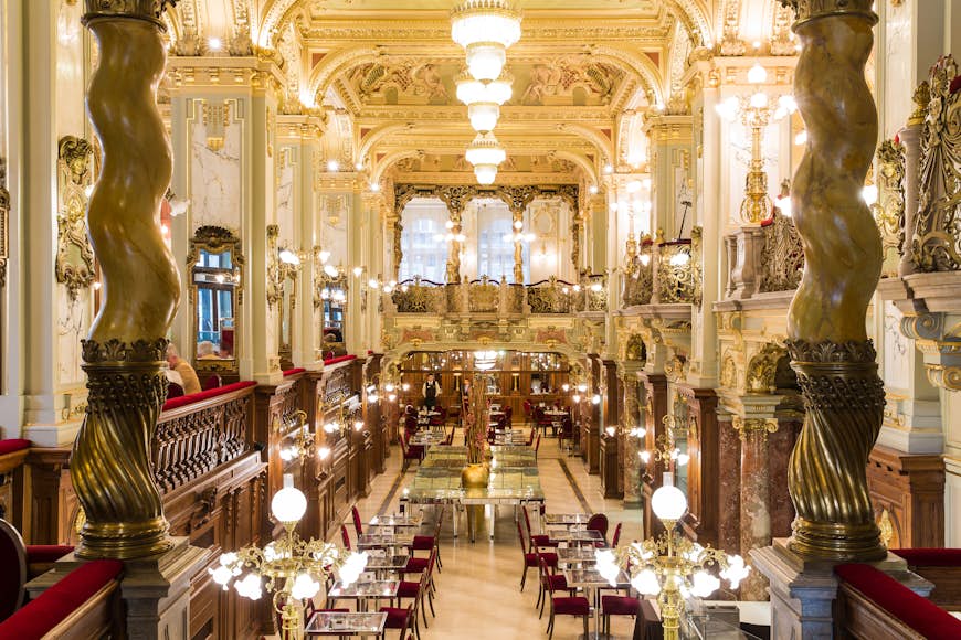 The wildly ornate interior of the New York Cafe in Budapest. There are marble columns, golden light fixtures and plush crimson chairs 