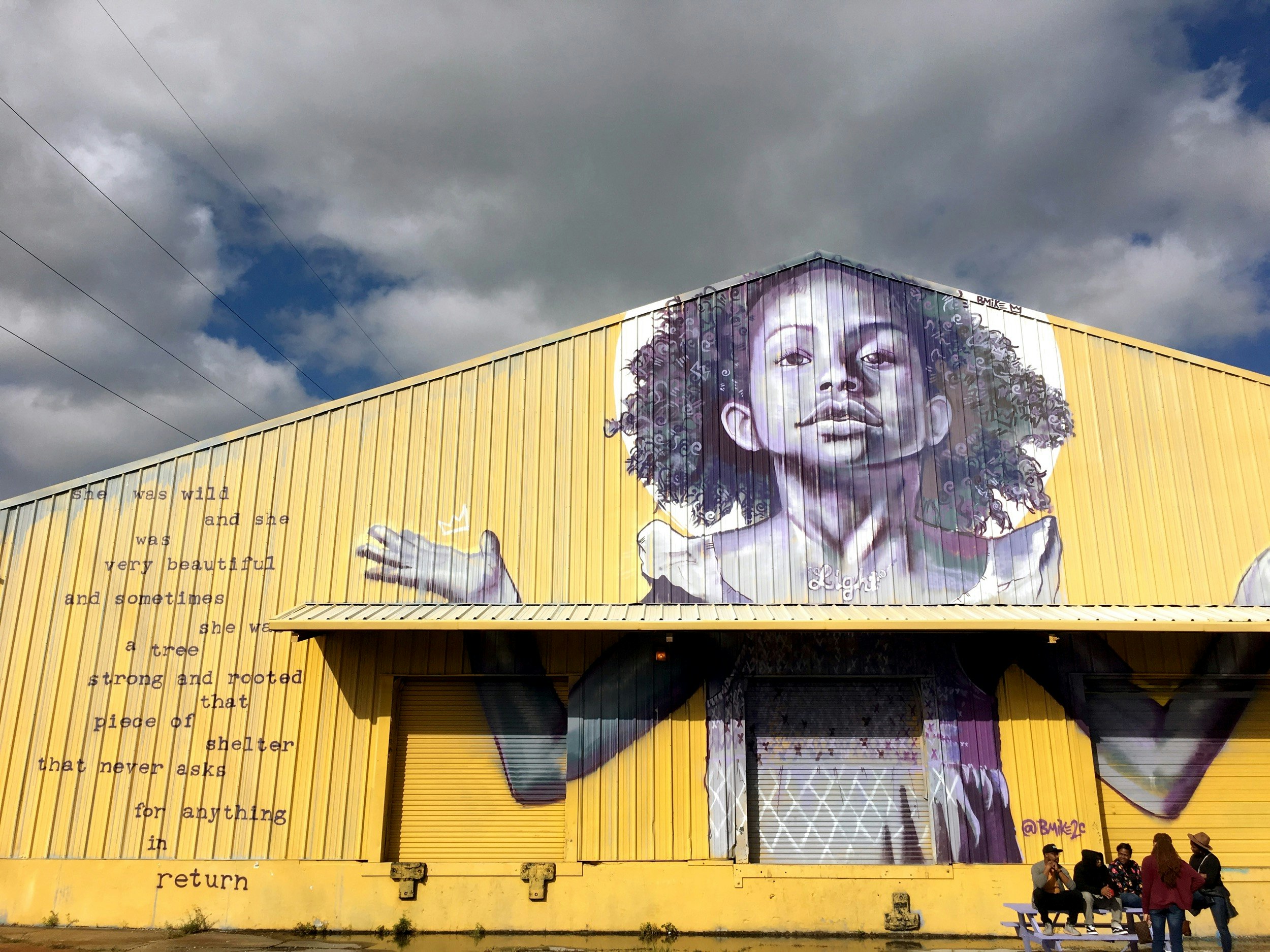 A yellow building with poetry painted on the left and a black and white image of a black girl with her arms raised in New Orleans
