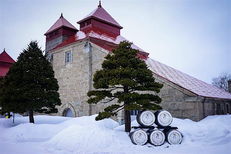 The Nikka Distillery at Yoichi in Hokkaido; we see a grey stone building with red towers, surrounded by snow with pine trees in front of it.