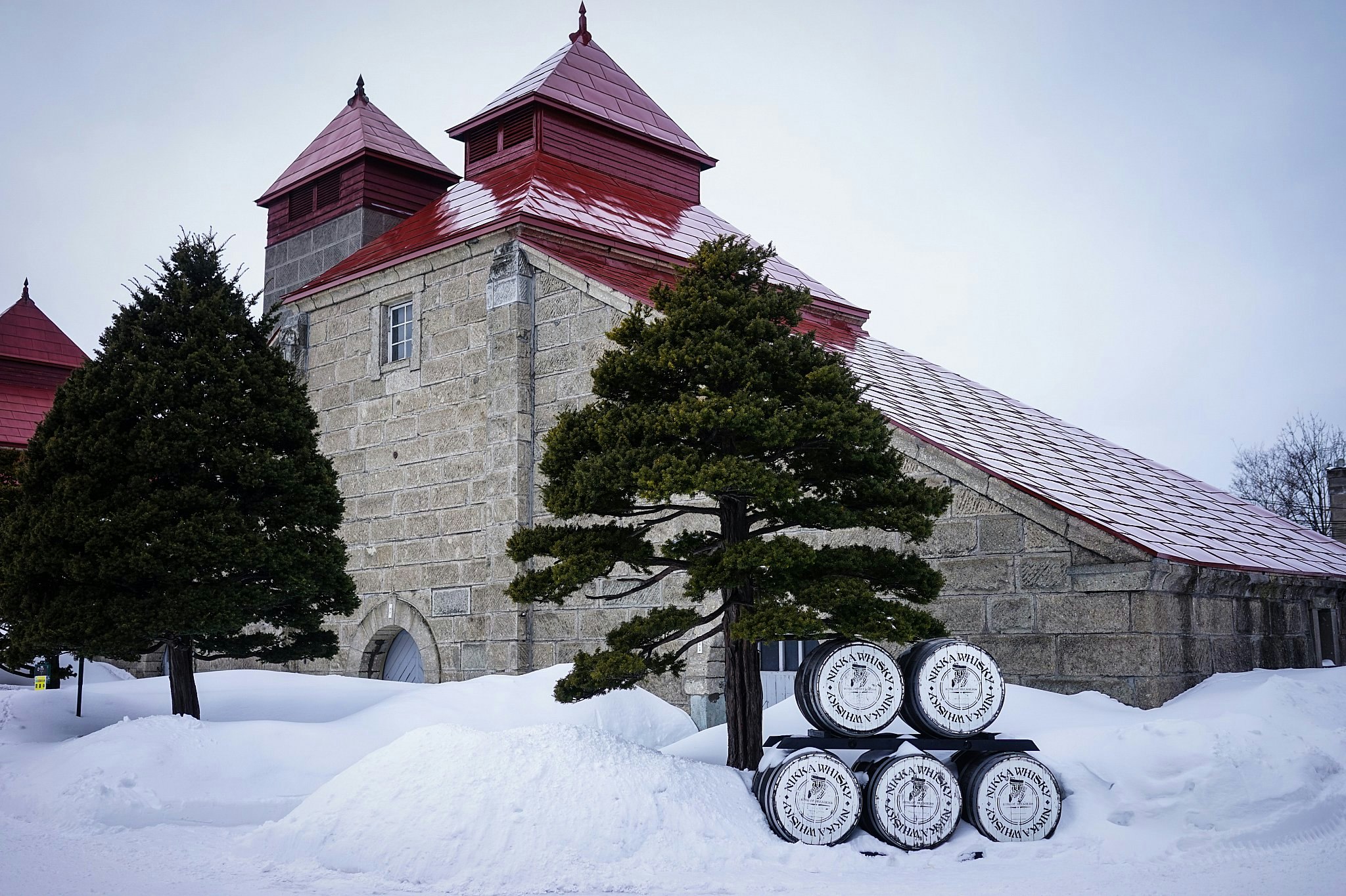 The Nikka Distillery at Yoichi in Hokkaido; we see a grey stone building with red towers, surrounded by snow with pine trees in front of it.