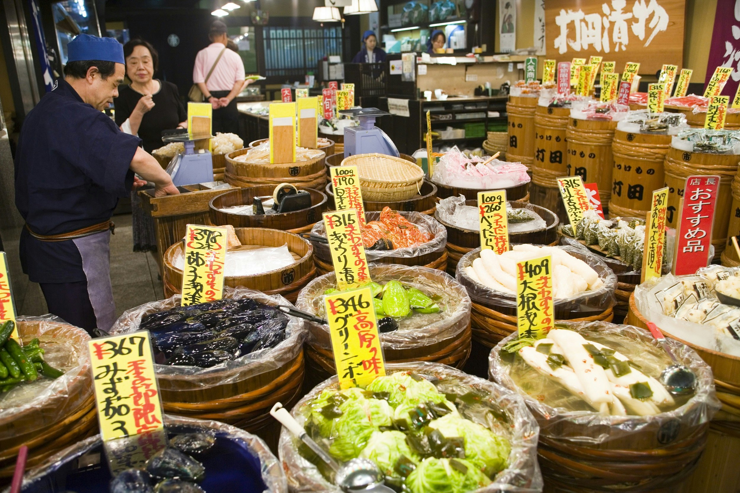 Numerous wooden tubs are filled with vegetables at the Nishiki Market; yellow labels with black-and-red Japanese writing denote the cost and contents of each.