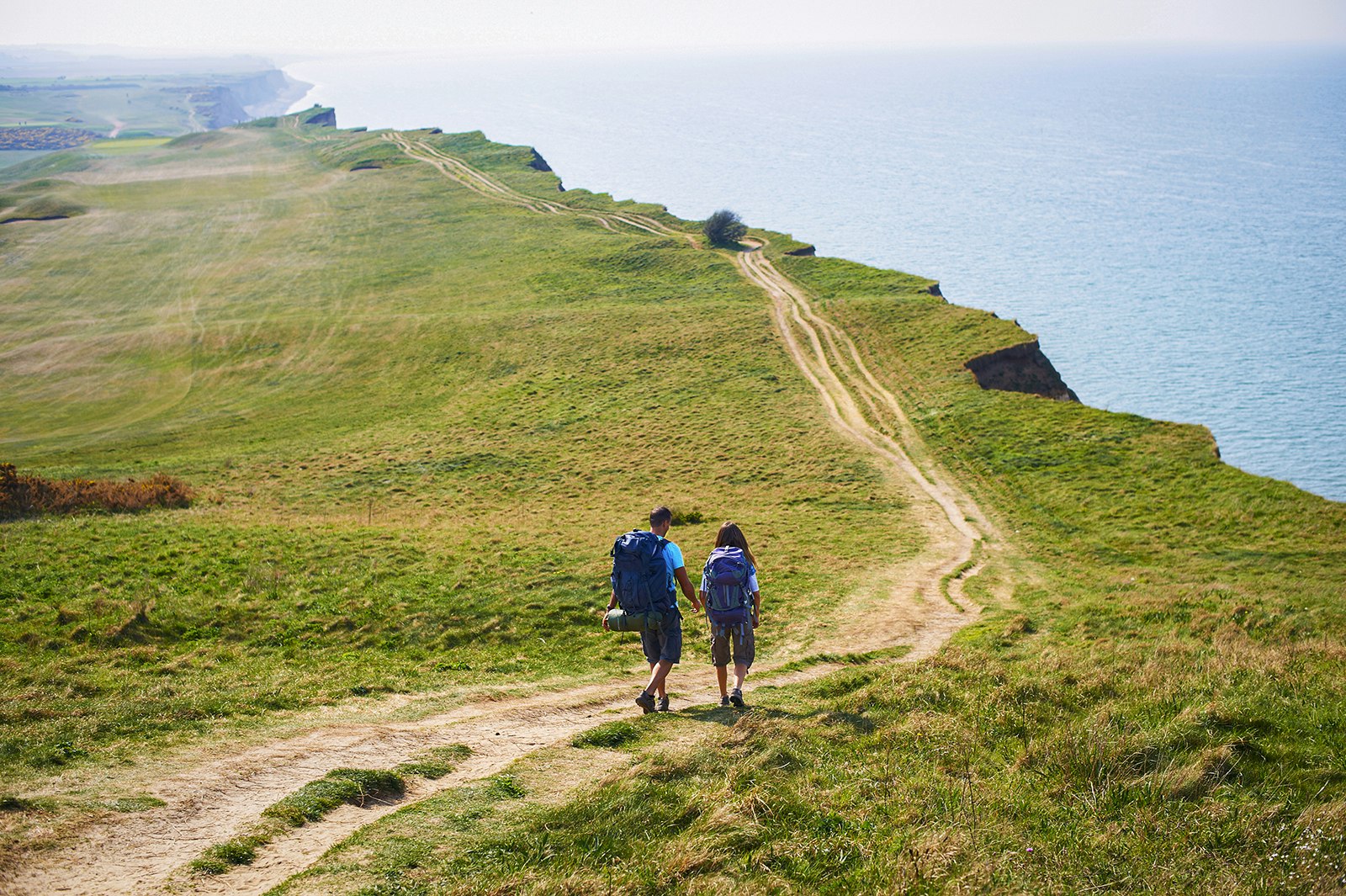 Two backpackers walk down a dirt path that cuts through a green field and hugs a cliff line. Norfolk Coast Path, England.