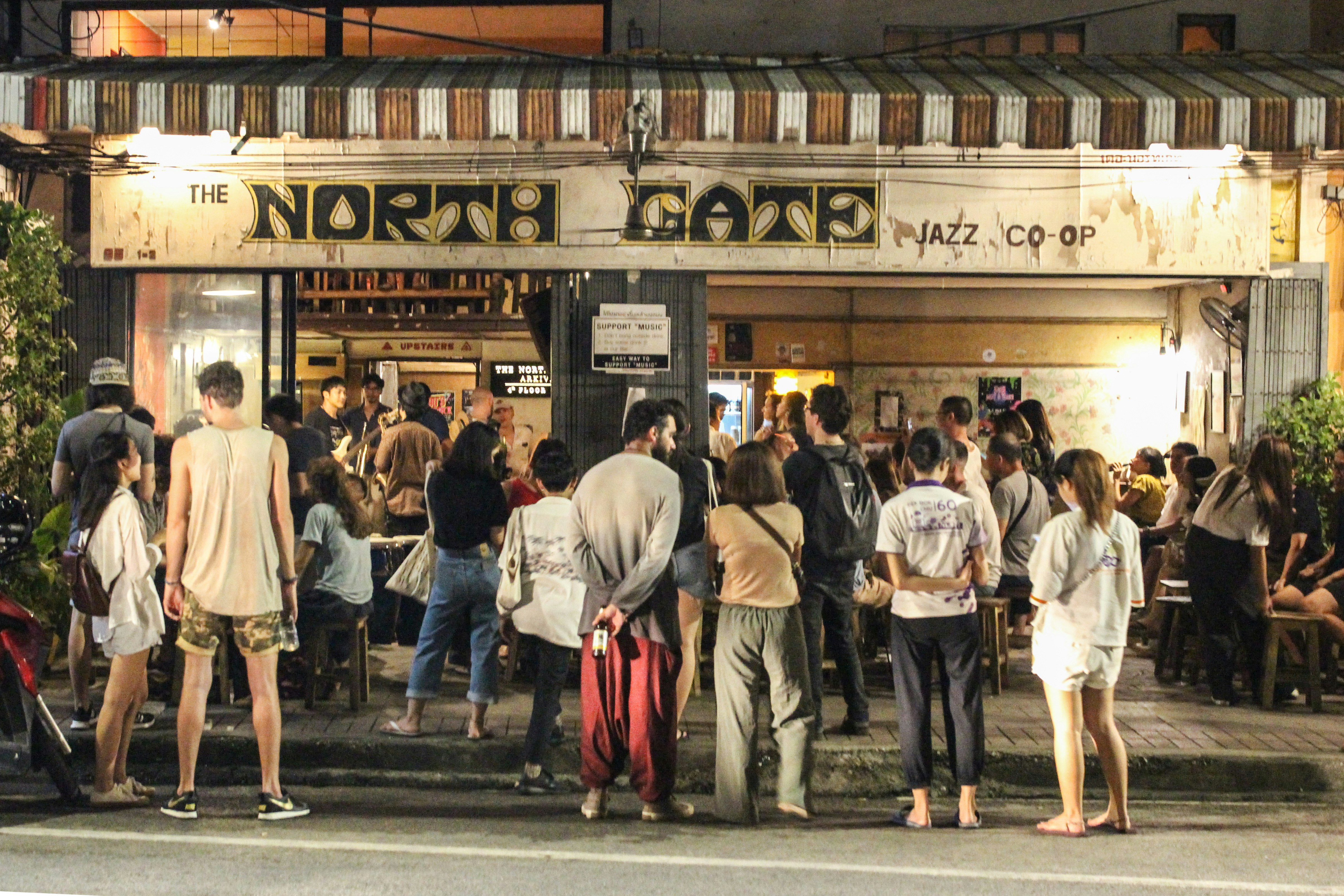 A large crowd of people gather at the North Jazz Co-Op bar to hear live music. The crowds spill from the inside of the bar onto the pavement outside.