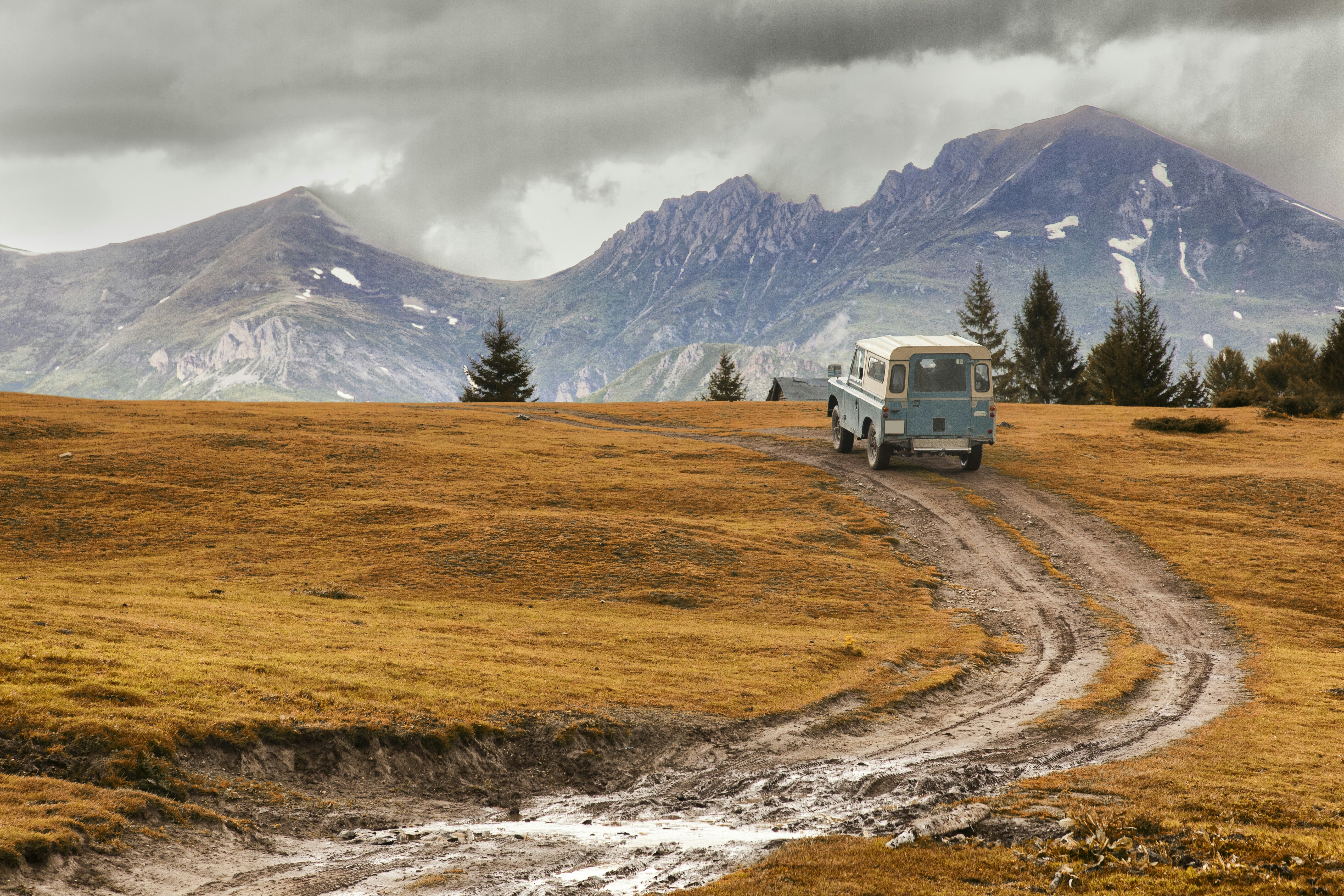 A light blue van dives up a muddle hill in an open field. In the background there are large snow-capped mountains; North Macedonia Best in Travel