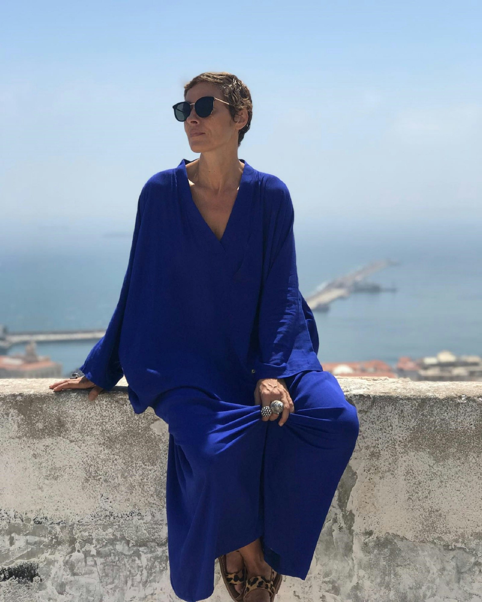 Fashion designer Norya sits atop a wall in a long blue dress, wearing sunglasses, large silver rings and leopard-print sandals. In the background is the sea in the distance.