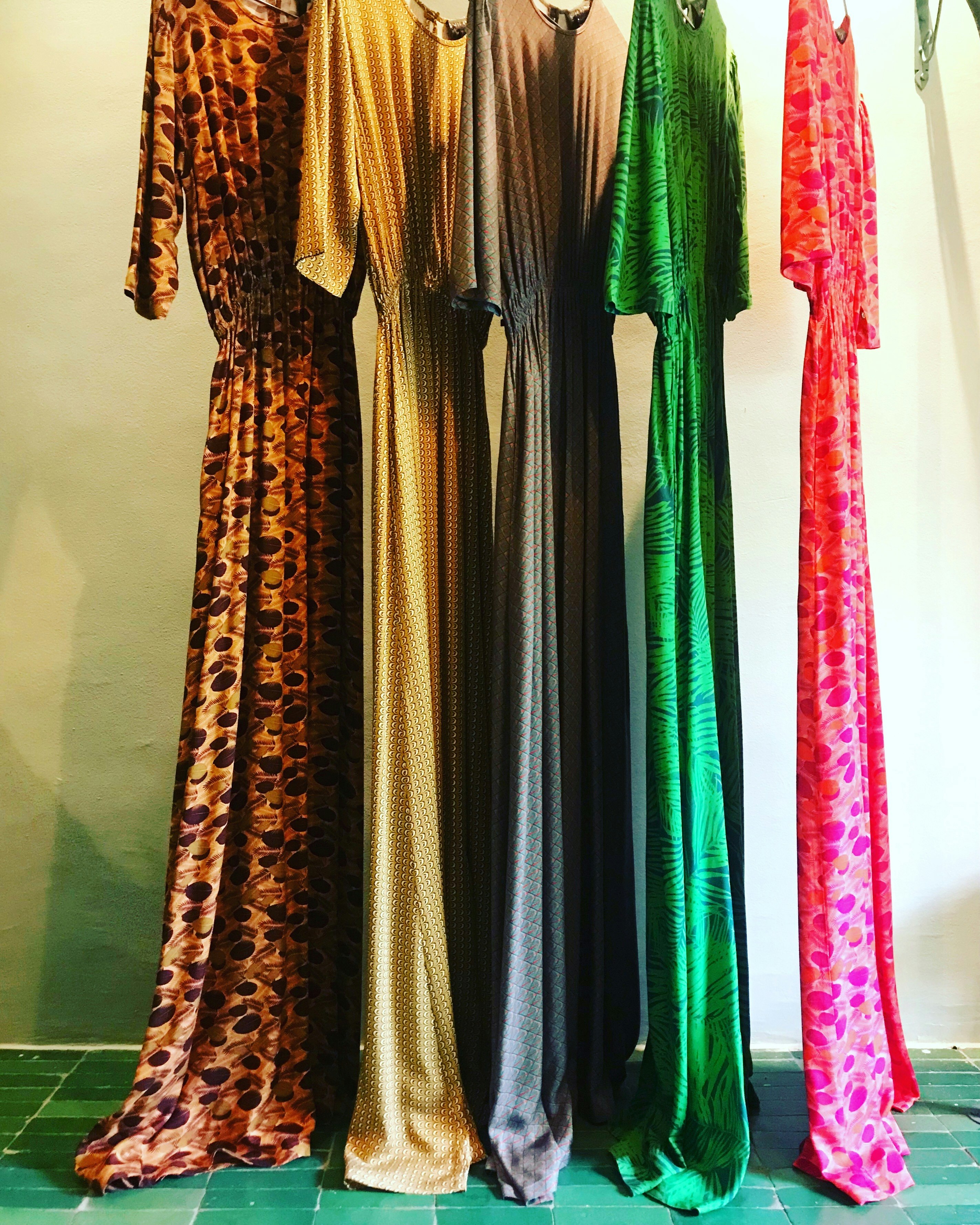 Five long silk dress of varied colours (green, gold, purple, red, silver, bronze) hang gracefully from hangers in front of a whitewashed wall.