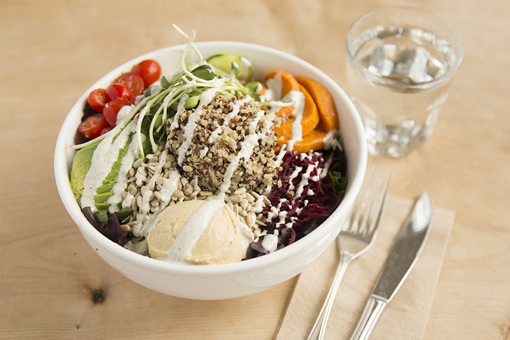 A bowl filled with quinoa, avocado, sweet potatoes, tomatoes and red cabbage sits next to a glass of water and a fork and knife.