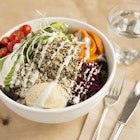 A bowl filled with quinoa, avocado, sweet potatoes, tomatoes and red cabbage sits next to a glass of water and a fork and knife.