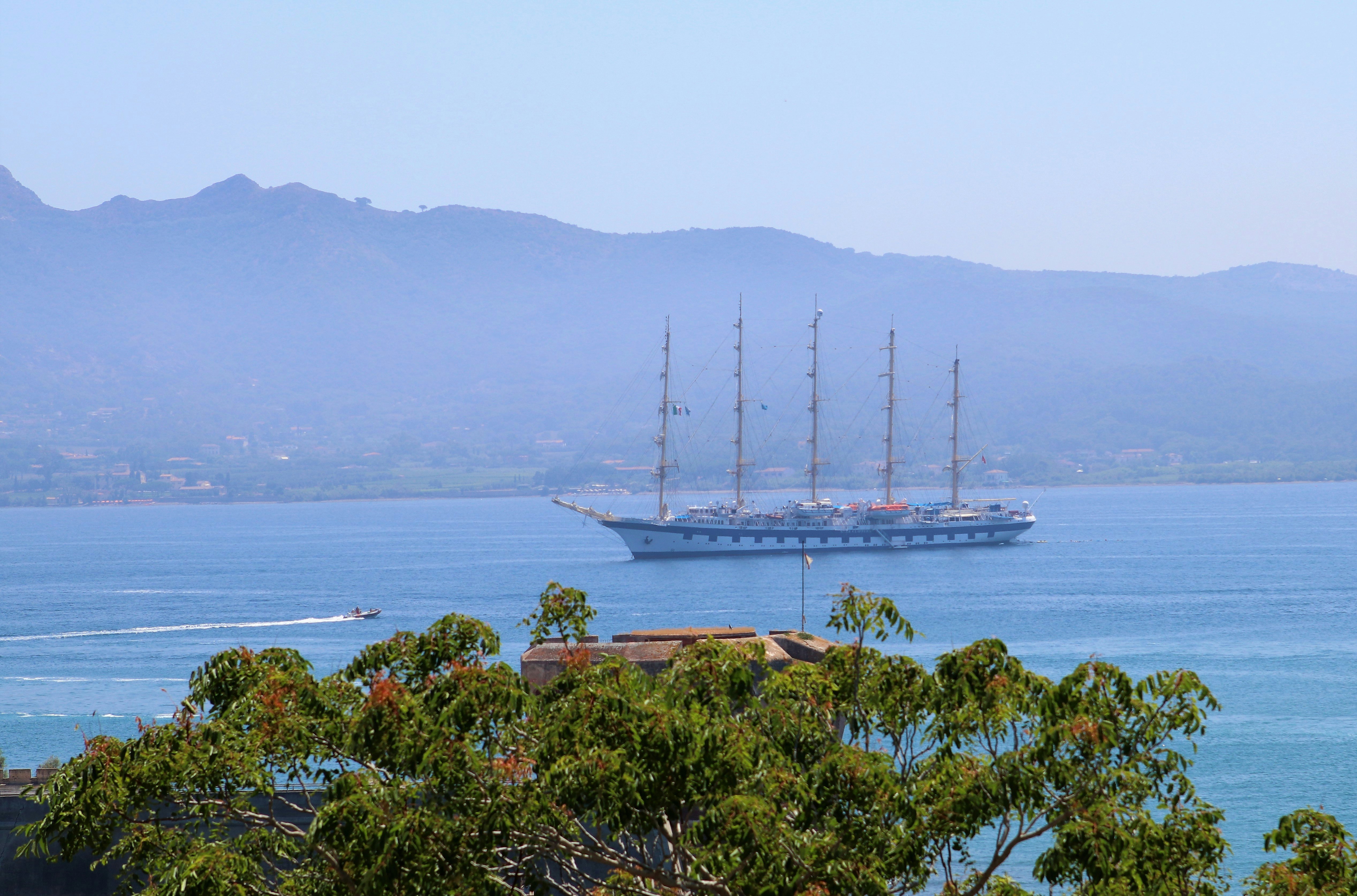 A large schooner glides across a body of water as a smaller speeds past. In the background is a small range of mountains; nude cruise 