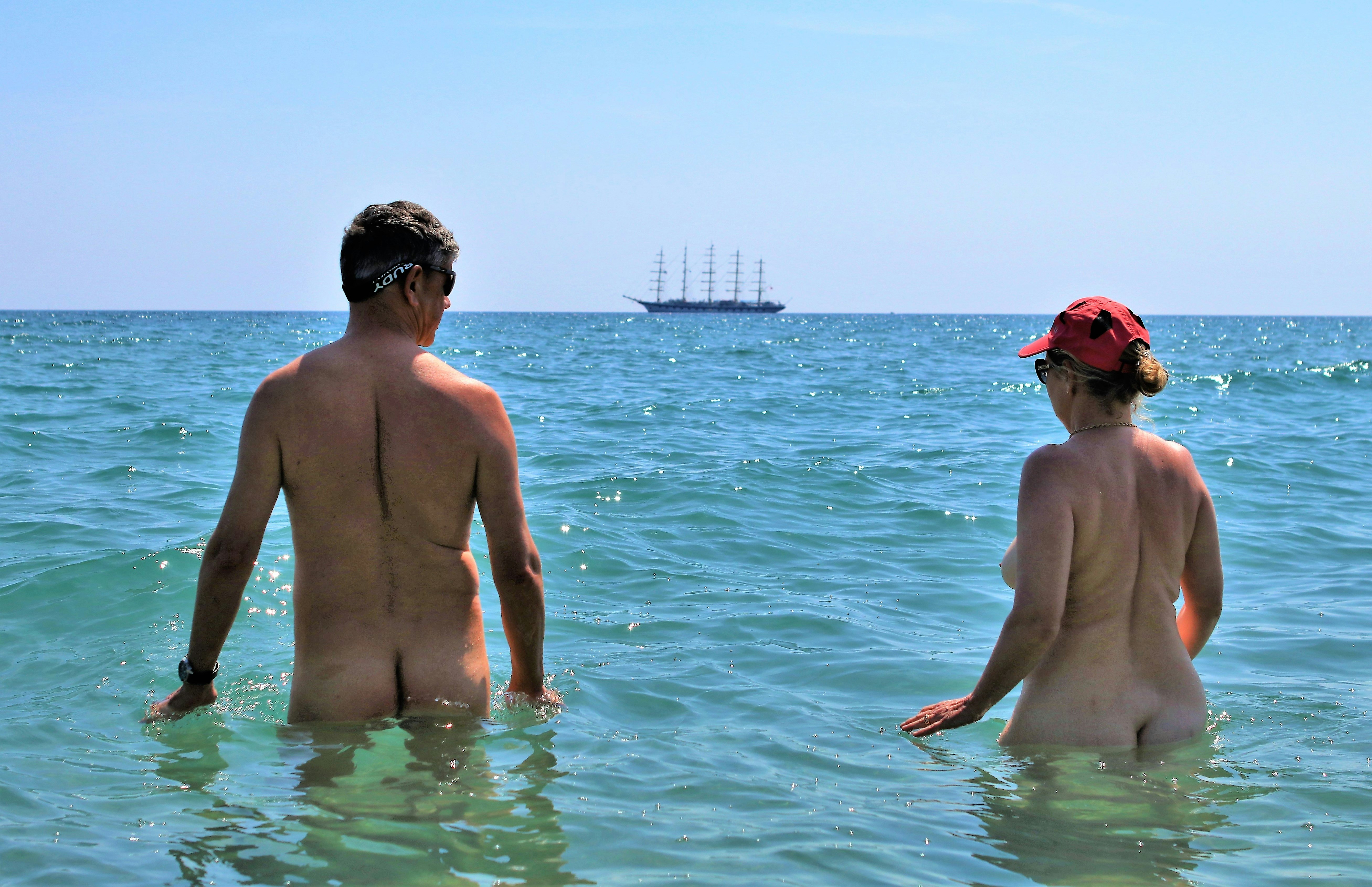 A couple wades through waist-high ocean water completely nude, though the man is wearing shades and the lady is wearing a baseball caps and sunglasses. There is a large schooner boat in the distance.