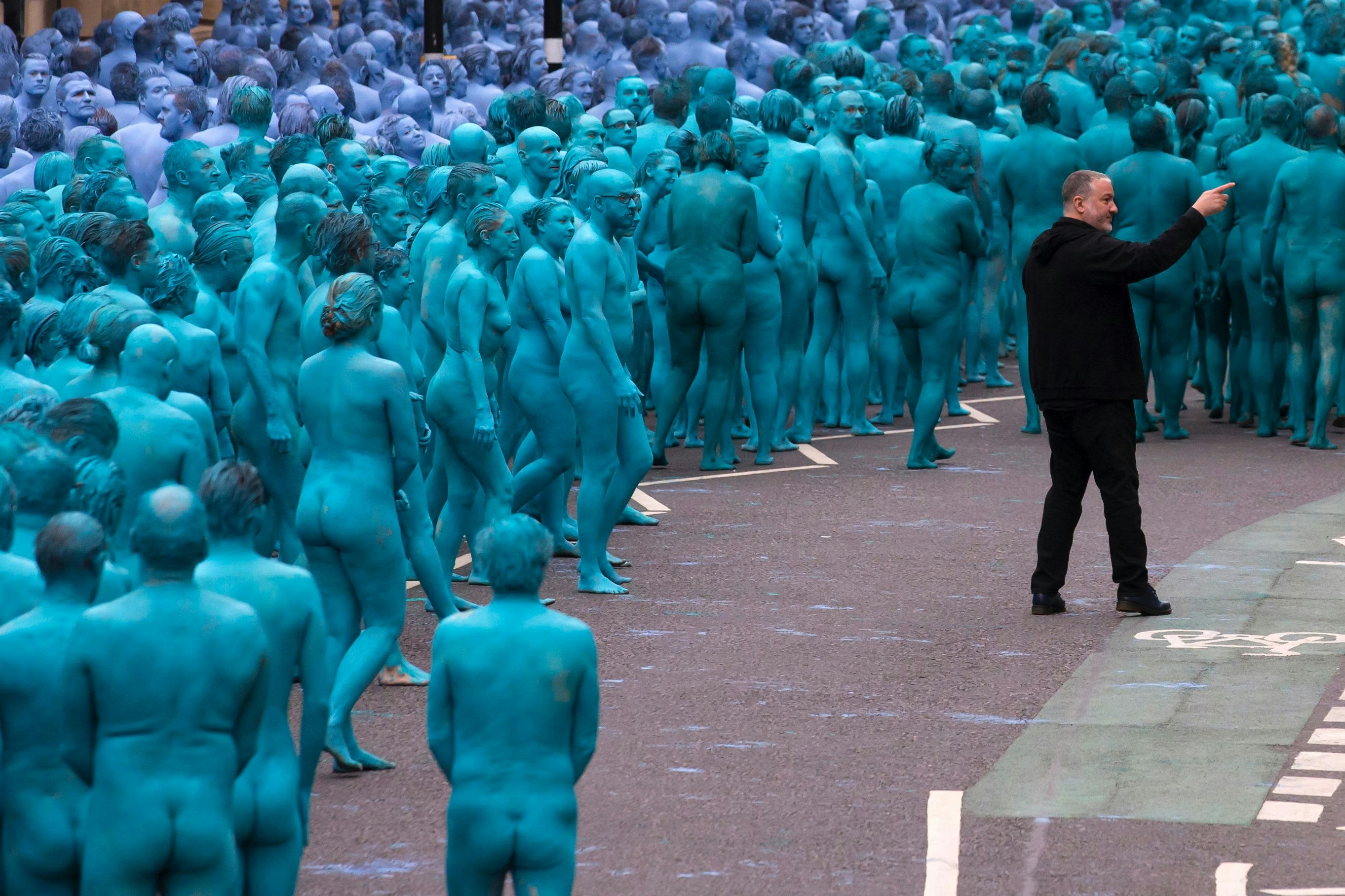 A large group of people fully naked and painted in varying shades of blue and purple stand on a city street as photographer Spencer Tunick points to someone off the image