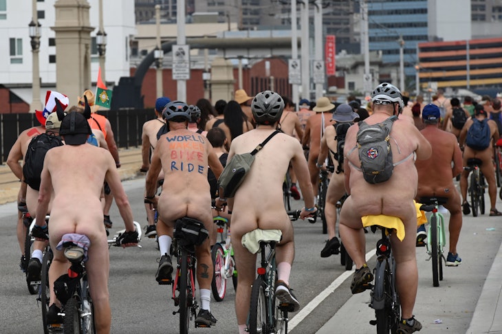 A group of cyclists ride through a city street in the nude. Some are wearing helmets and all are wearing socks and sneakers.