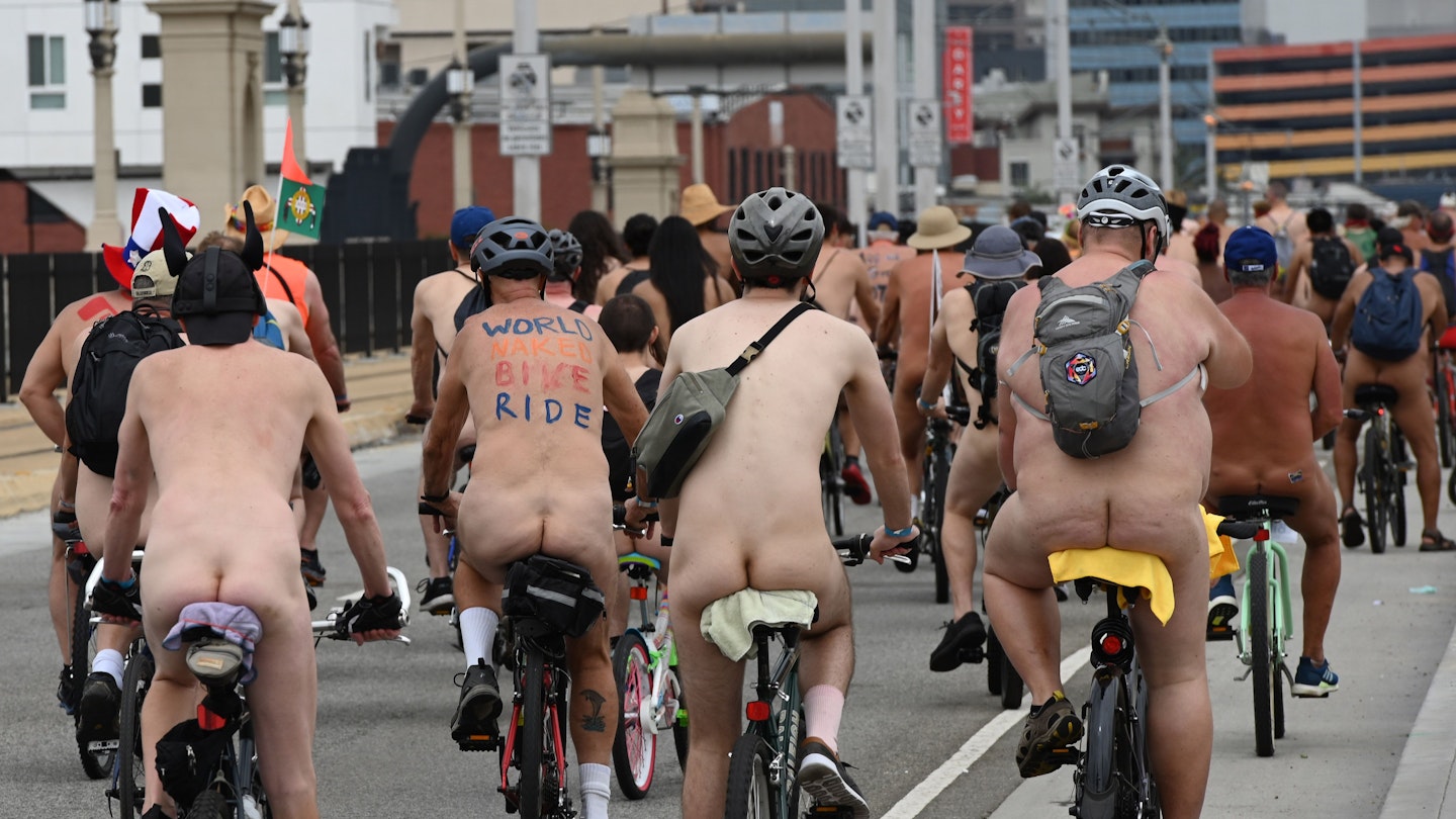 A group of cyclists ride through a city street in the nude. Some are wearing helmets and all are wearing socks and sneakers.