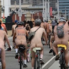 A group of cyclists ride through a city street in the nude. Some are wearing helmets and all are wearing socks and sneakers.
