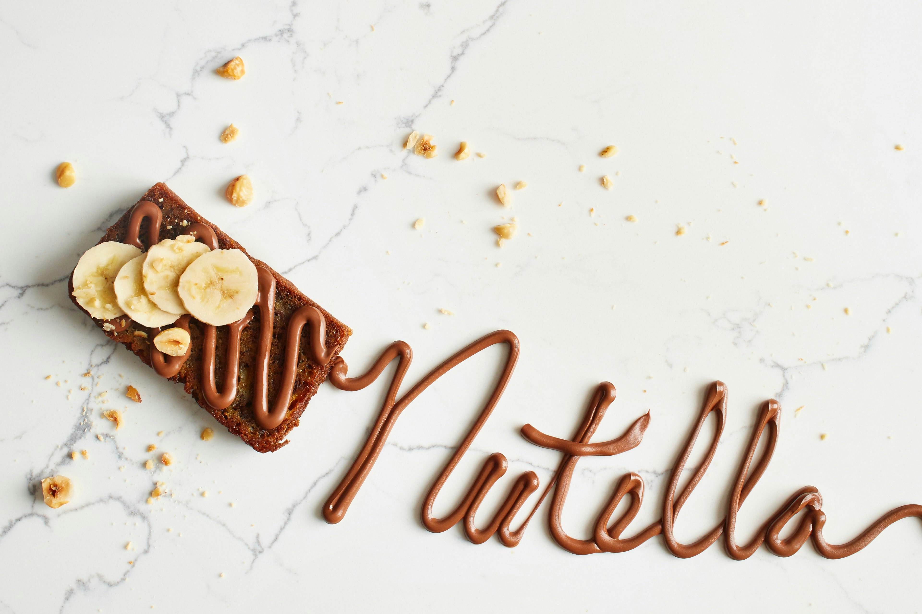 A piece of brioche topped with sliced bananas, hazelnuts, and Nutella, with the word "Nutella" written in Nutella