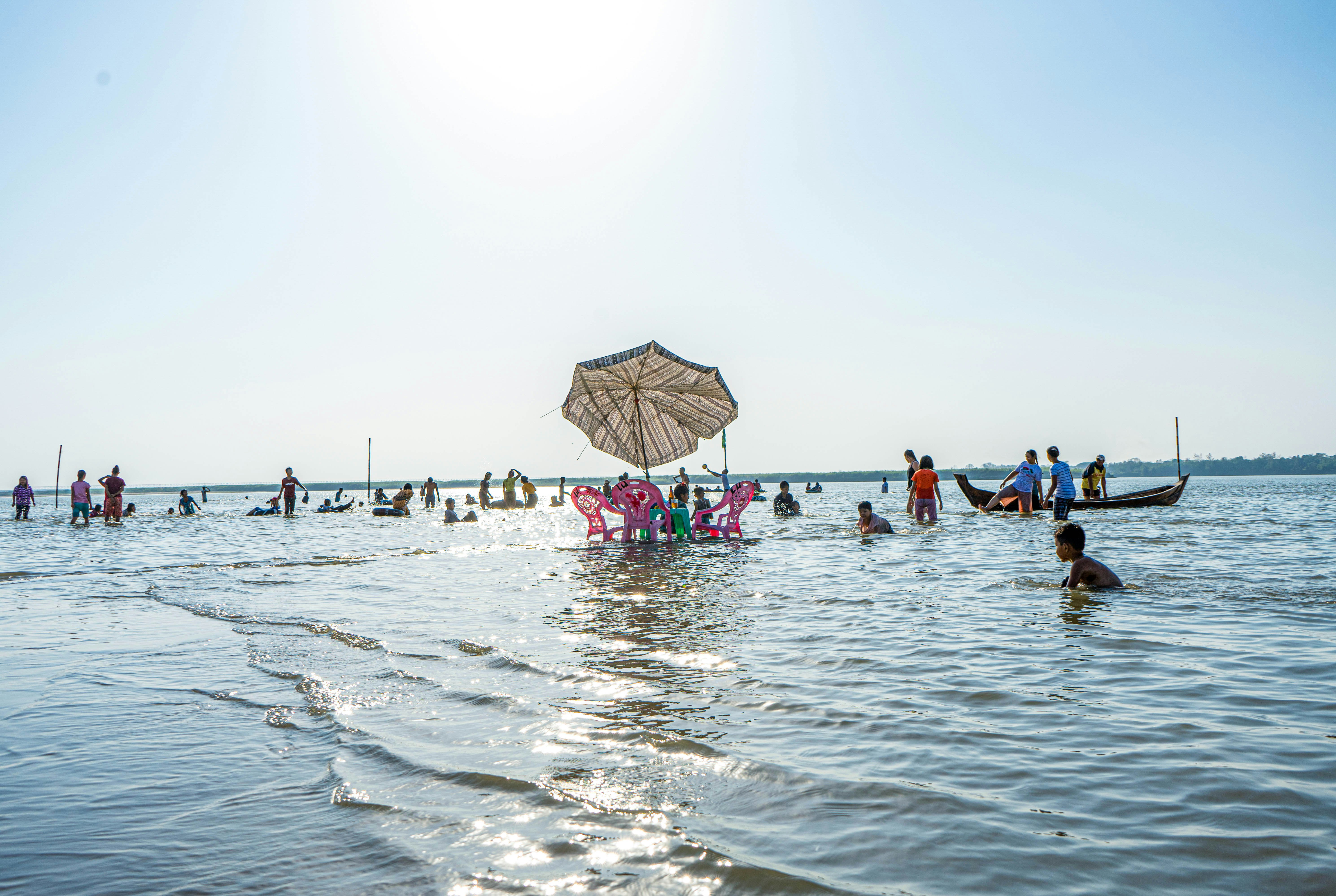 A table and three chairs are slowly submerged by the tide as it laps the beach in Myanmar. Many people are paddling in the shallow waters.