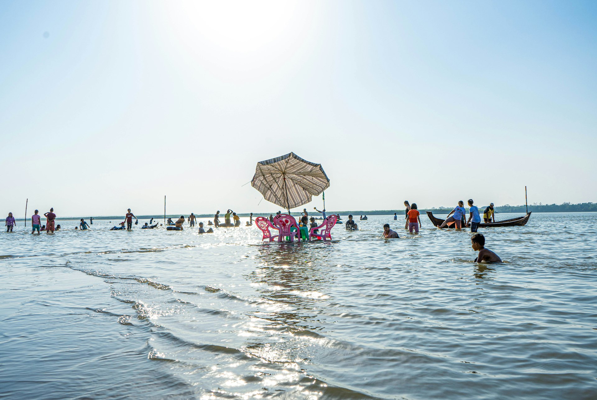A table and three chairs are slowly submerged by the tide as it laps the beach in Myanmar. Many people are paddling in the shallow waters.