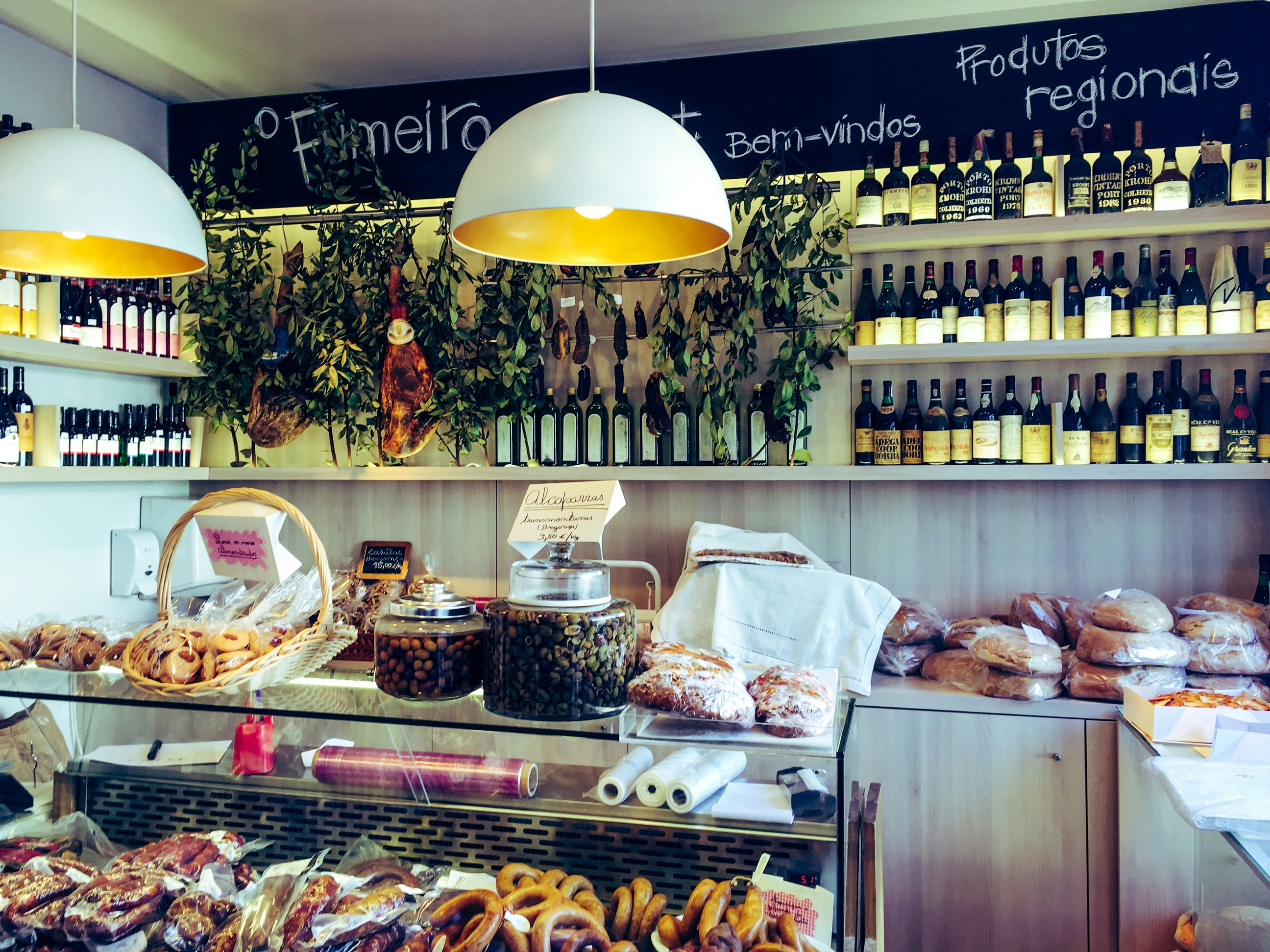 A glass display counter is filled with baked goods and topped with jars and baskets of pastries and olives, while the wall behind is lined with bottles of oil, vinegar and wine.