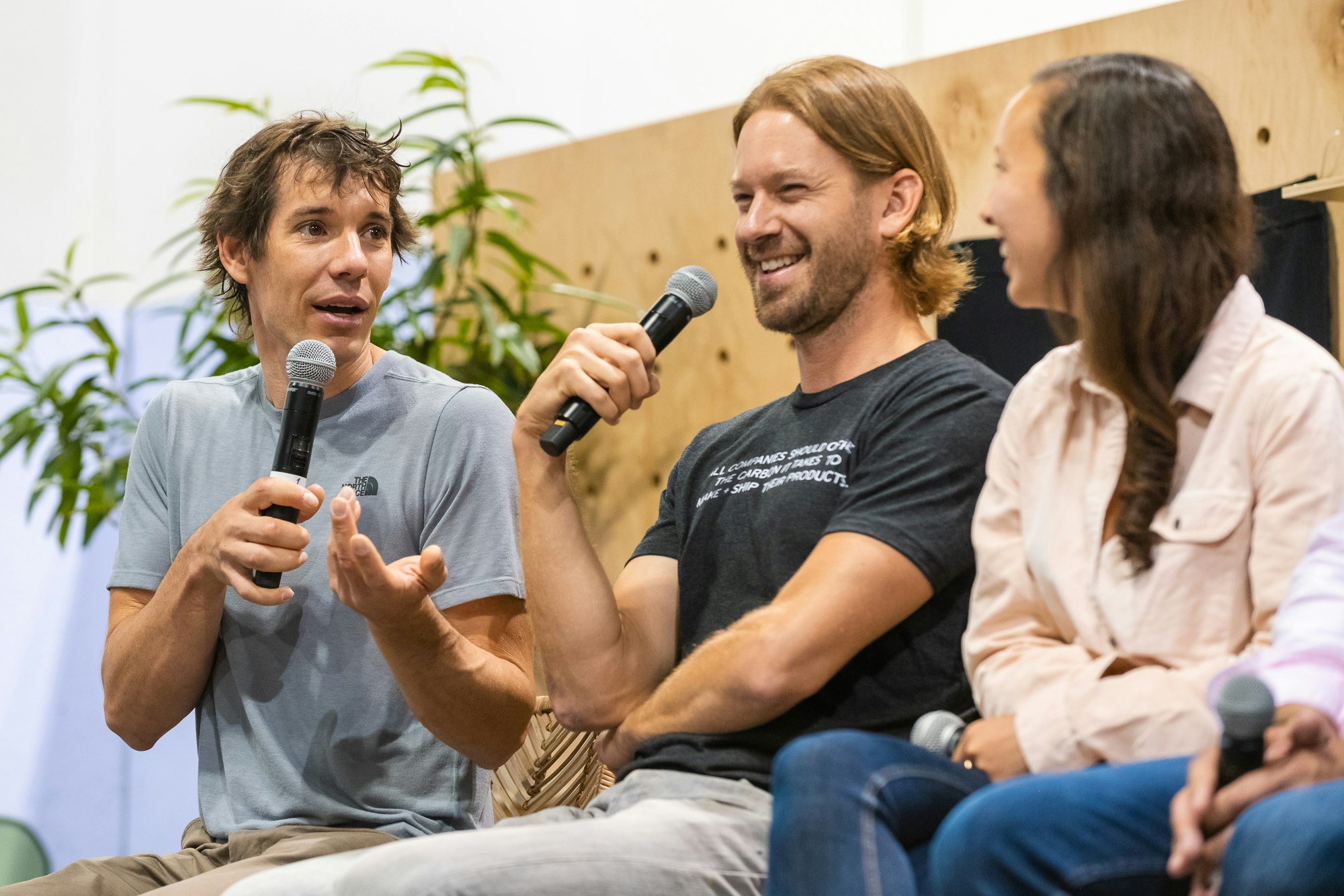 Alex Honnold, with short, shaggy brown hair and brown eyes, holds a microphone and gestures with his left hand while speaking on a panel with Peter Dering in a black t-shirt with shite text on the chest and long strawberry brown hair tucked behind one ear as he laughs and Hana Kajimura in a pink blouse with long loose brown hair turns away from the viewer towards her fellow panelists.  