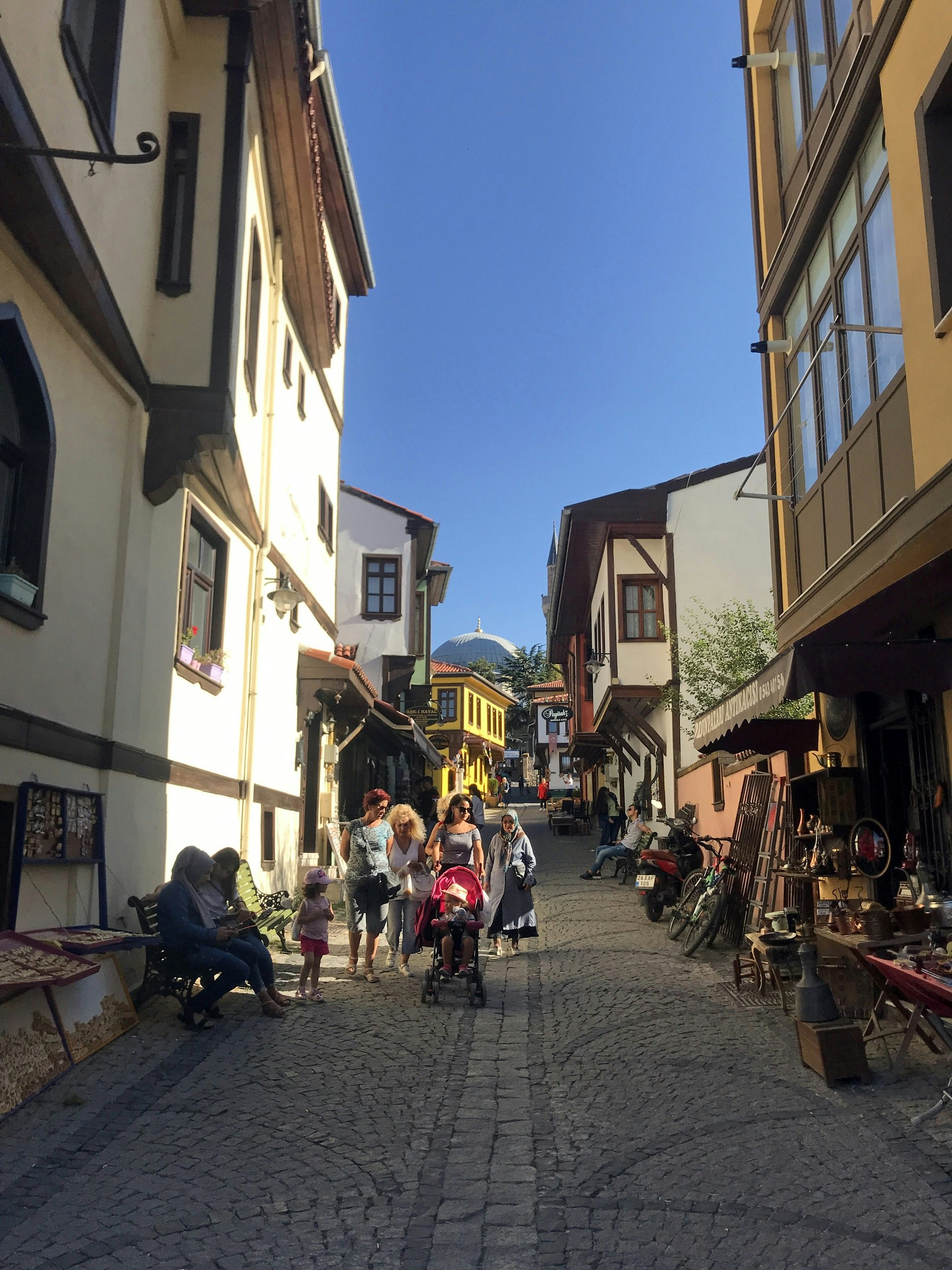 A cobbled street lined with traditional houses, some with shops or stalls alongside the road. A family strolls down the hill with a toddler and young child in a pram