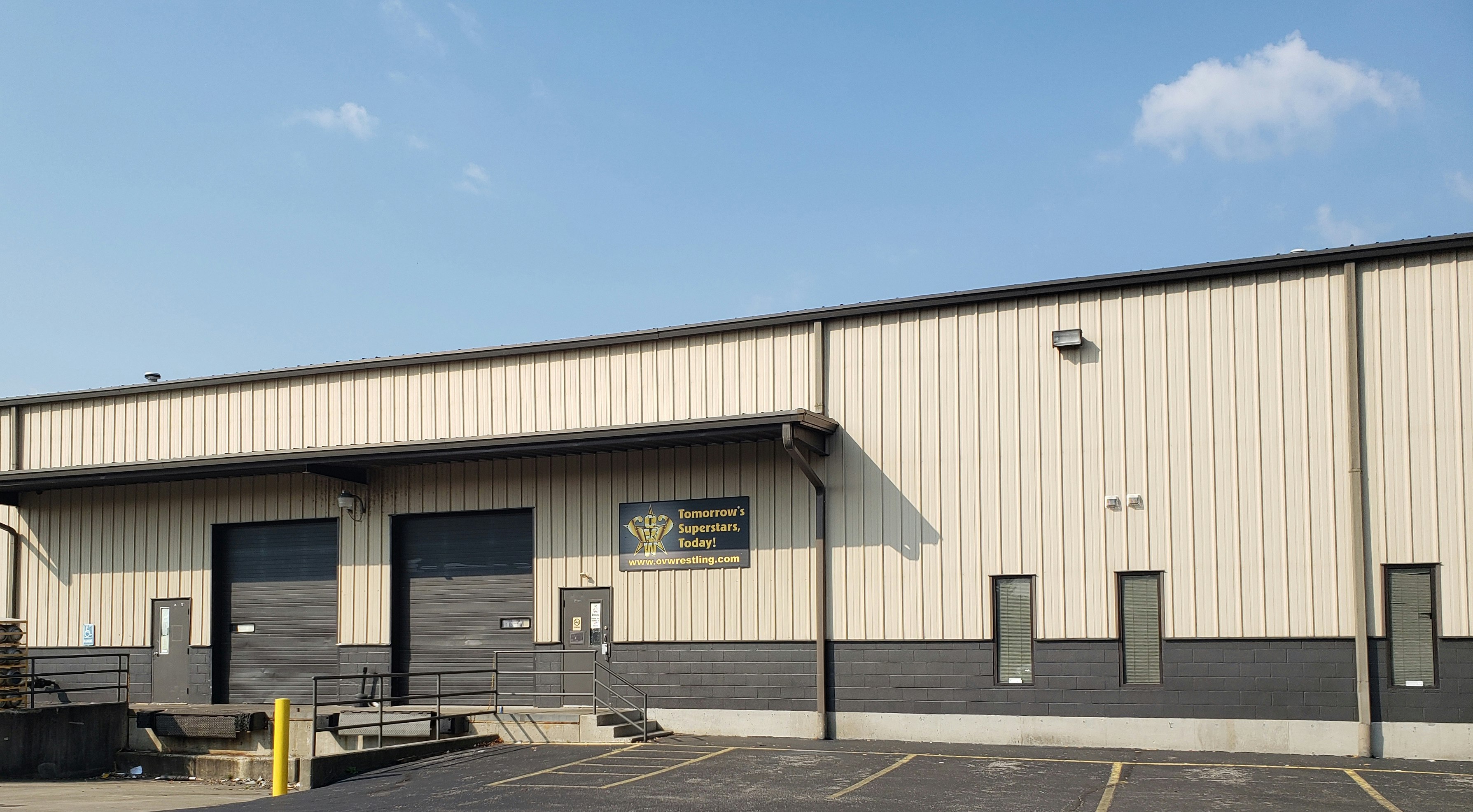 A tan corrugated metal exterior of the Ohio Valley Wrestling trade school has an industrial appearance, complete with two large, dark brown garage doors over loading docks. A black sign by the door with the OVW logo has bright yellow letters that read Tomorrow's Superstars, Today!  