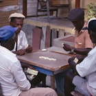 Four older gentlemen sit around a table playing dominoes in front of a Barbados rum shop