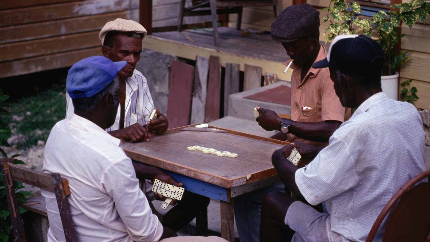 Four older gentlemen sit around a table playing dominoes in front of a Barbados rum shop