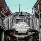 The words "Ole Smoky Moonshine" are painted on a dark wood sign attached to a wooden deck. "Old Smoky Holler" is spelled out in lights above the wood deck.  
