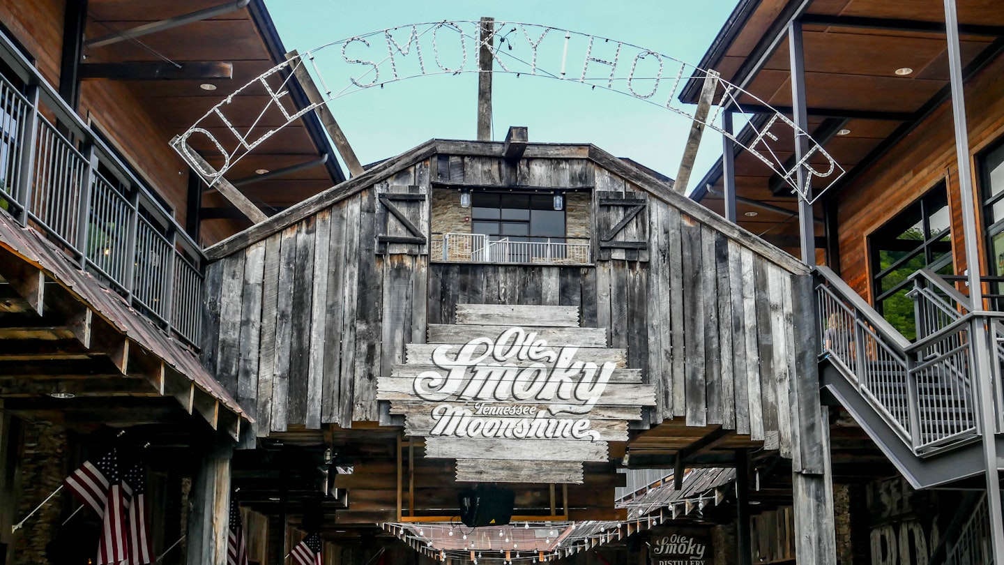 The words "Ole Smoky Moonshine" are painted on a dark wood sign attached to a wooden deck. "Old Smoky Holler" is spelled out in lights above the wood deck.  