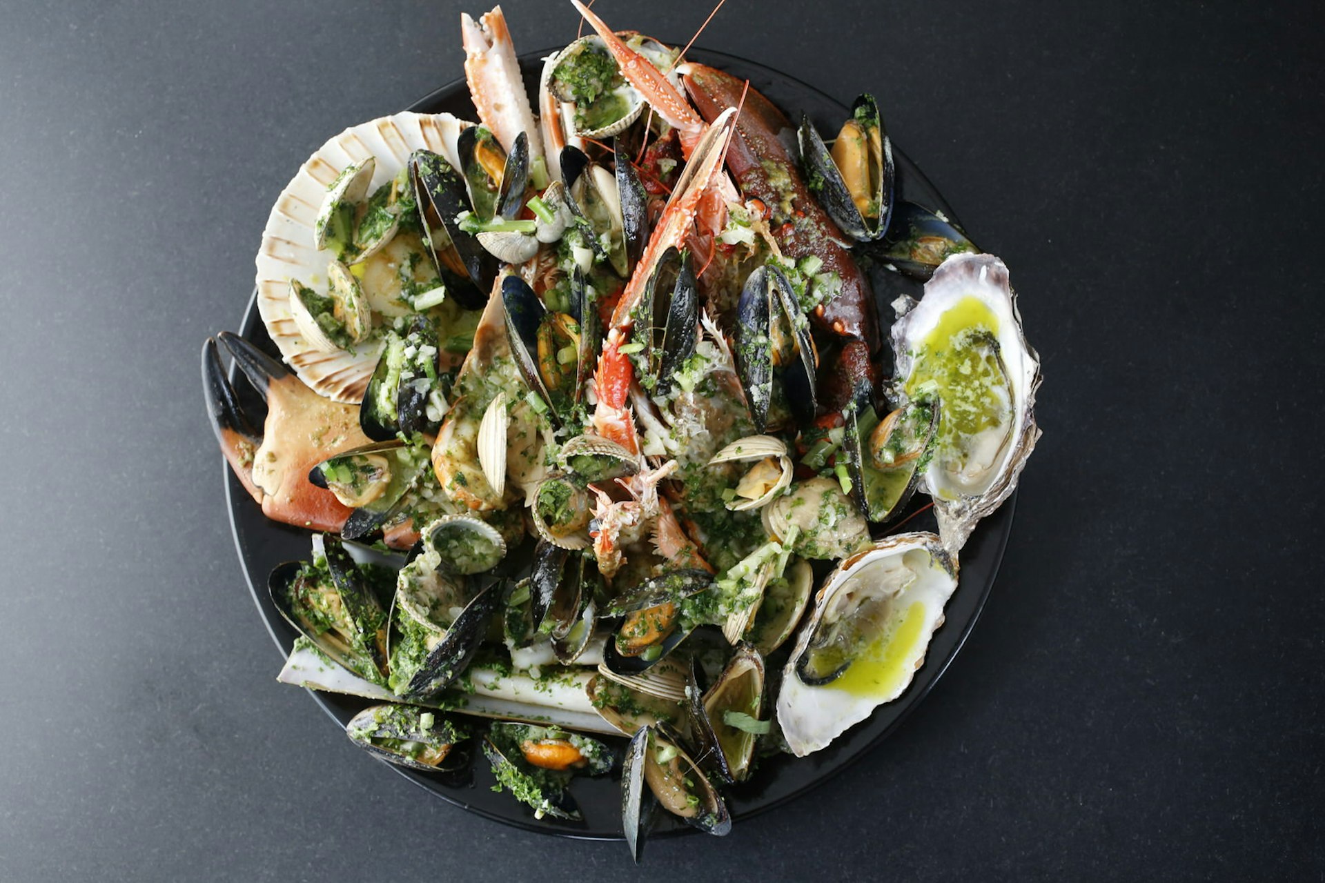 A black plate on a black table stuffed with fresh seafood, served at Ondine restaurant in Edinburgh. The plate features oysters, lobster, mussels, clams, scallops and crab.