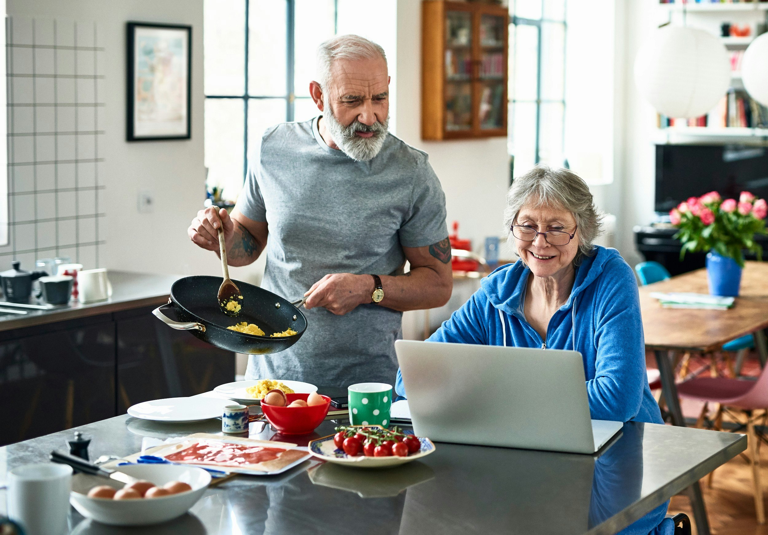 A senior man and woman cooking food and looking at a laptop