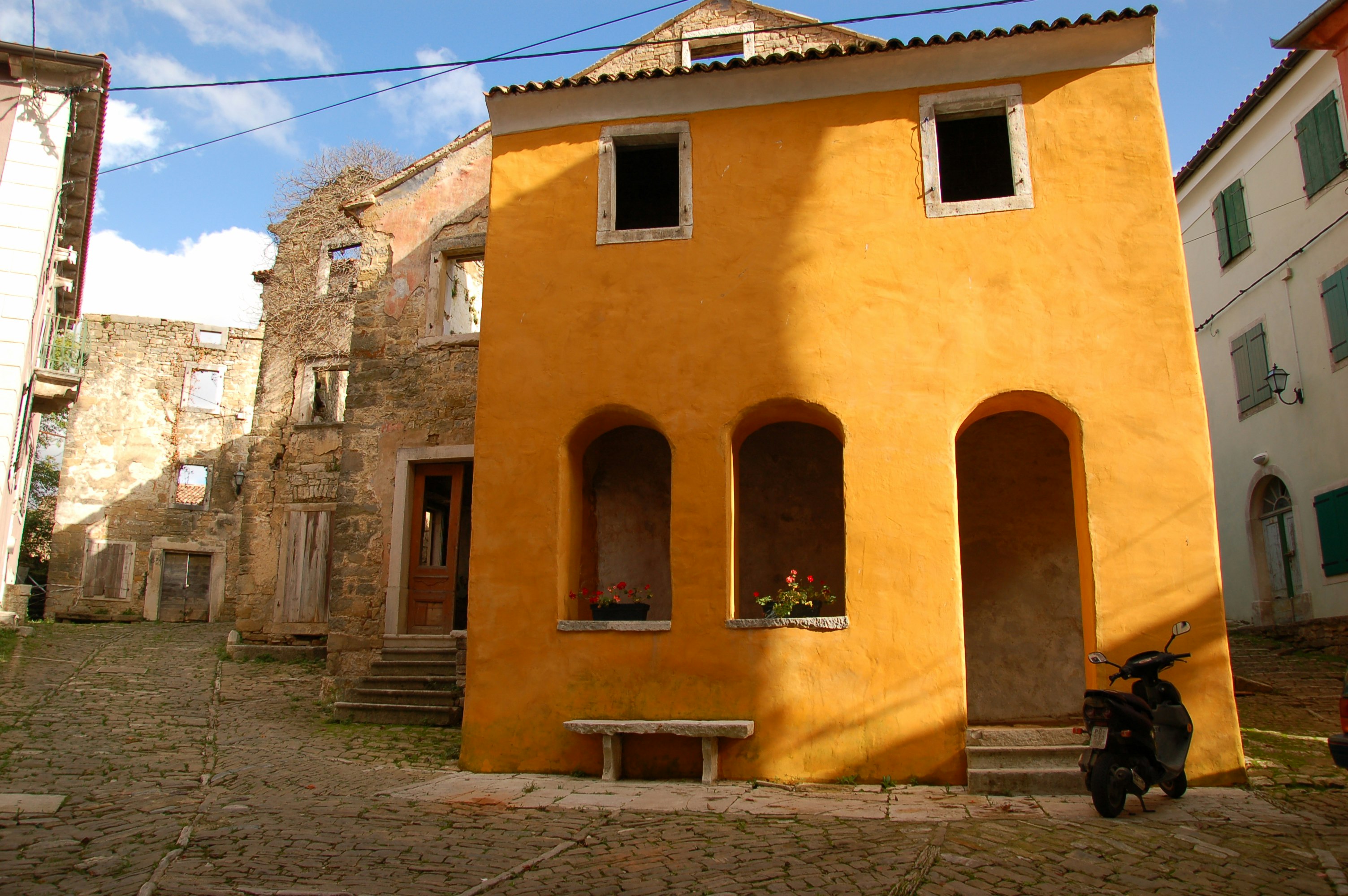 An orange stucco building with arched windows and doors on the ground floor and square windows rimmed in white sills on the upper floor sits in the winding medieval streets of Oprtalj. Behind the orange building are empty ruins of other similar stone houses. In the foreground a motorcycle is parked  