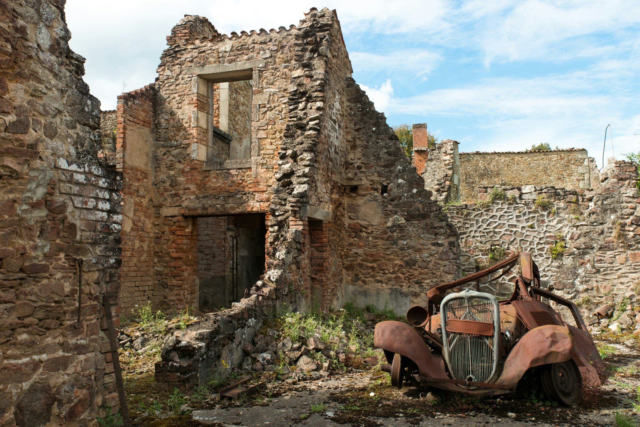 A rusted car lies in the ruins of a house in Oradour-Sur Glane in France, as the sun shines down.