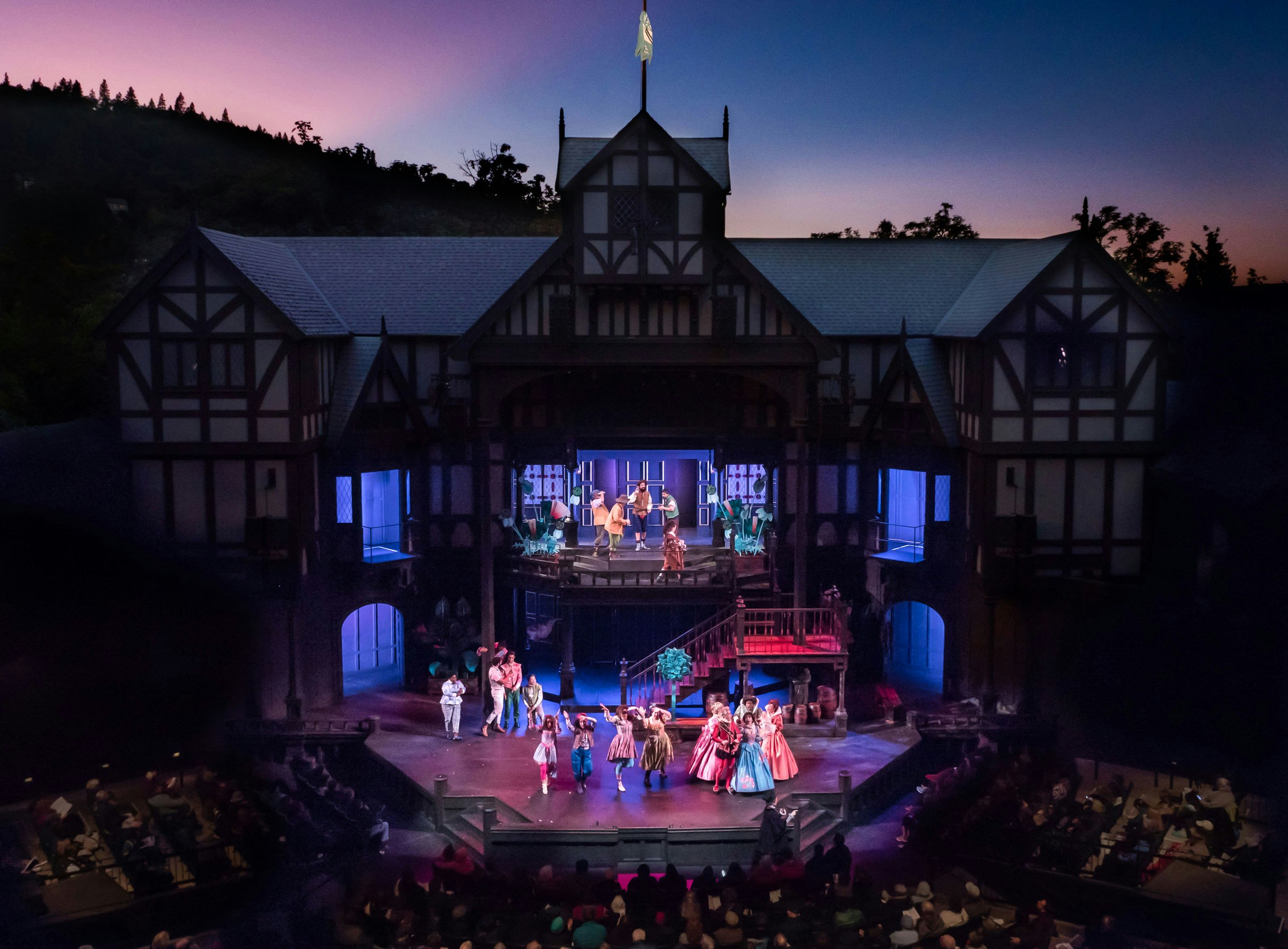 Oregon Shakespeare Festival performers onstage in open-air theatre against a pink sunset.